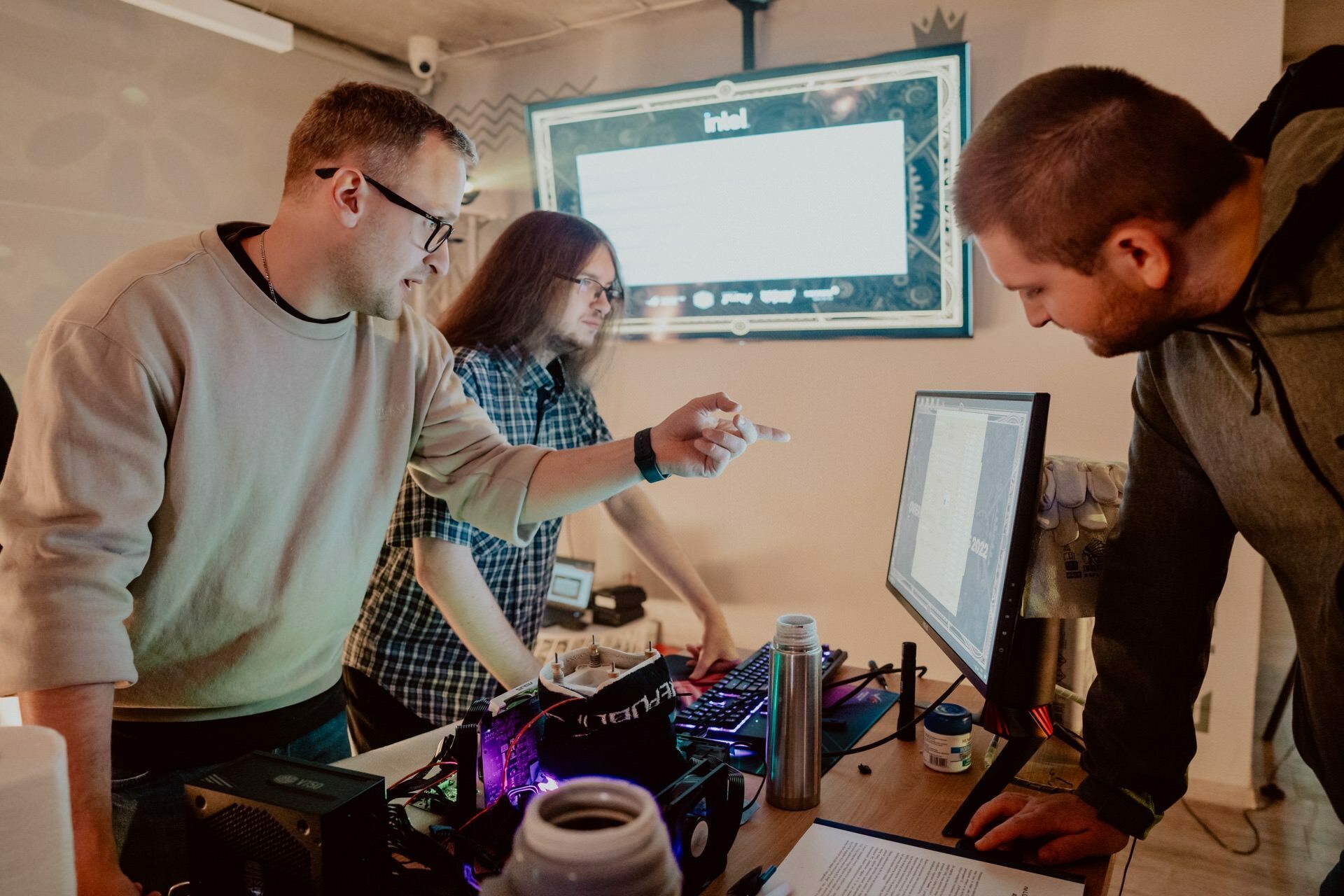 Three men gather around a desk with computer equipment. One man in a light-colored sweater points to the computer monitor, while the other two watch intently. Behind them on the wall is a large monitor displaying a presentation on which an in-depth photo report of events is being recorded.  