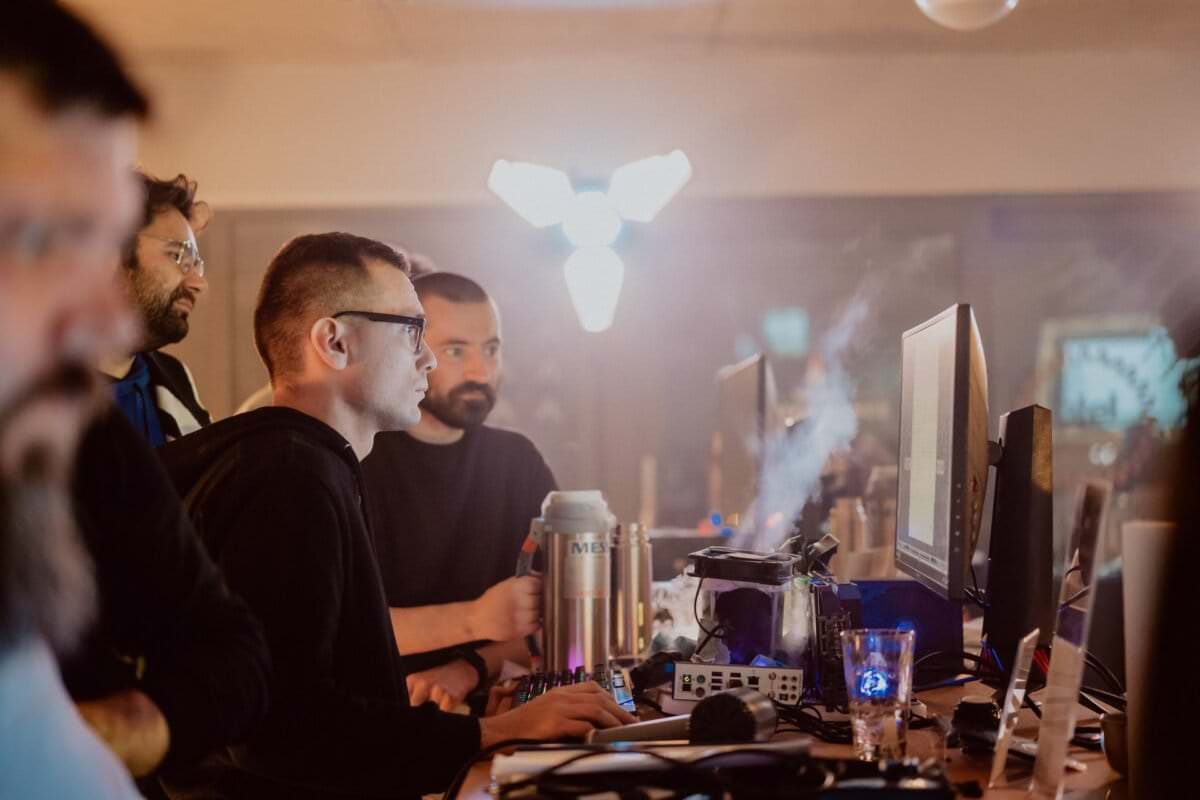 In a dimly lit room, a group of people are working in concentration at computer monitors. One person in the foreground sits and focuses on the screen, while others stand or sit nearby. The workspace is littered with equipment, and a bright lighting fixture hangs in the background, capturing the essence of event photography.  