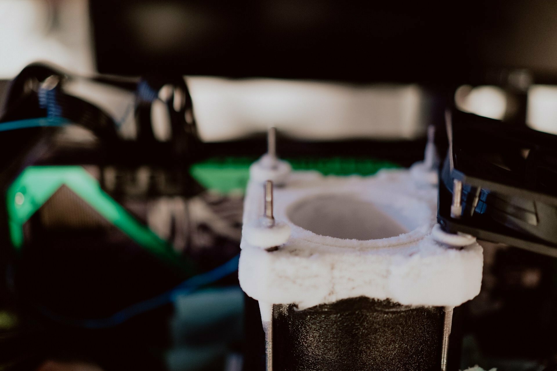 A close-up of the 3D printer in action. The image focuses on the printer extruding a bright filament, creating a cylindrical object. In the background, somewhat out of focus, you can see various parts of the printer, including rods and wires - a striking shot by Marcin Krokowski.  