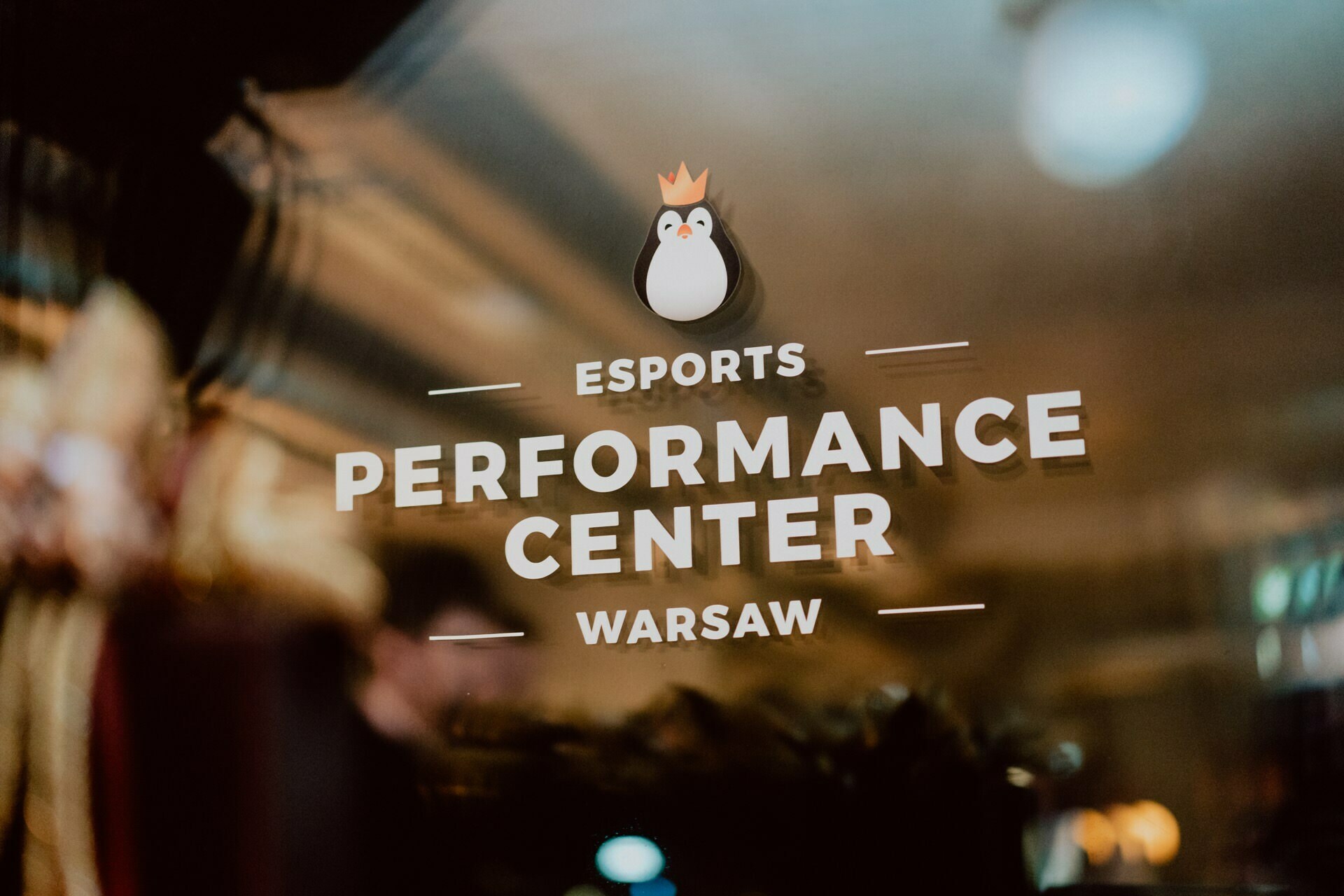 Glass door with a logo depicting a cartoon penguin wearing a crown. The door reads "Esports Performance Center Warsaw." The glass shows the reflection of the dimly lit interior, expertly captured by Marcin Krokowski, who specializes in event photography.  