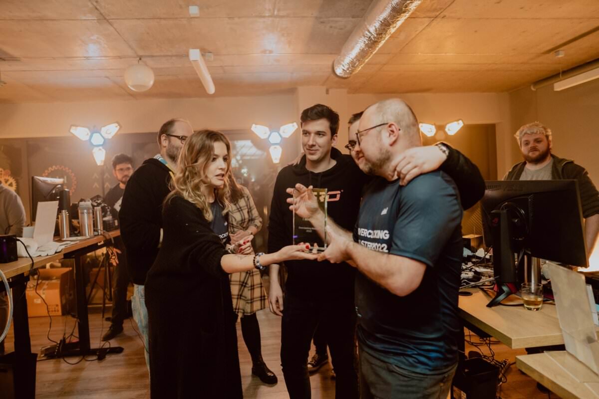 A group of people in a dimly lit room with industrial decor. One woman and two men in the foreground are engaged in conversation, while others stand in the background at desks and computer screens. The atmosphere seems casual and collaborative, reminiscent of an event photographer's photo essay.  