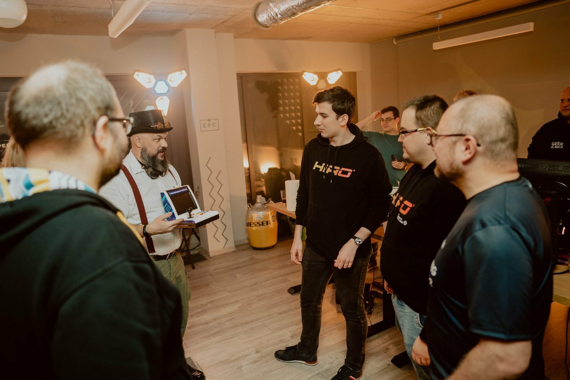 Six people, some wearing glasses and branded T-shirts, gather attentively in the well-lit office. Marcin Krokowski stands out in a hat and suspenders, talking to the group while holding open a laptop. This moment captures the dynamic photo coverage of the event.  