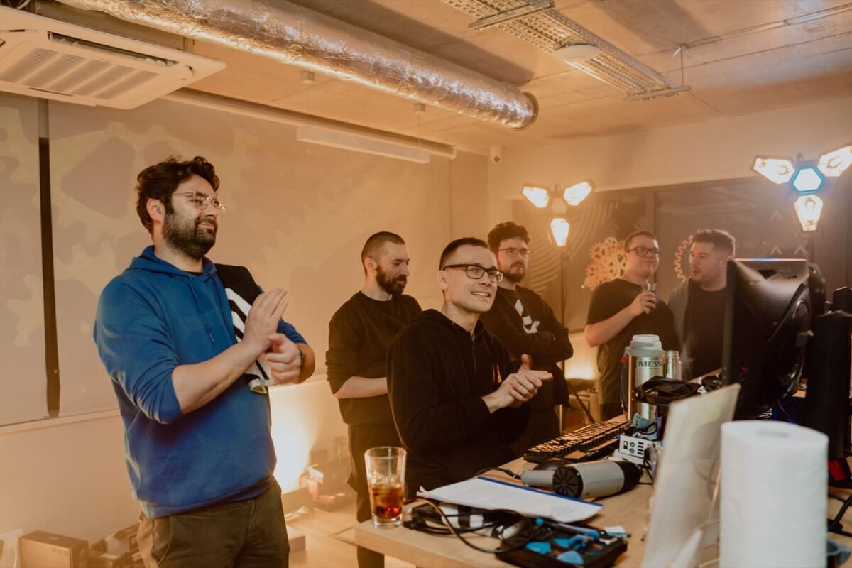 A group of six people in a modern office space, all looking toward computer screens. Some are standing, and someone is sitting with his hands on the keyboard. They appear to be collaborating, with various equipment and drinks on the table. The lighting is warm and atmospheric, creating the perfect atmosphere for photo opportunities of the events.   