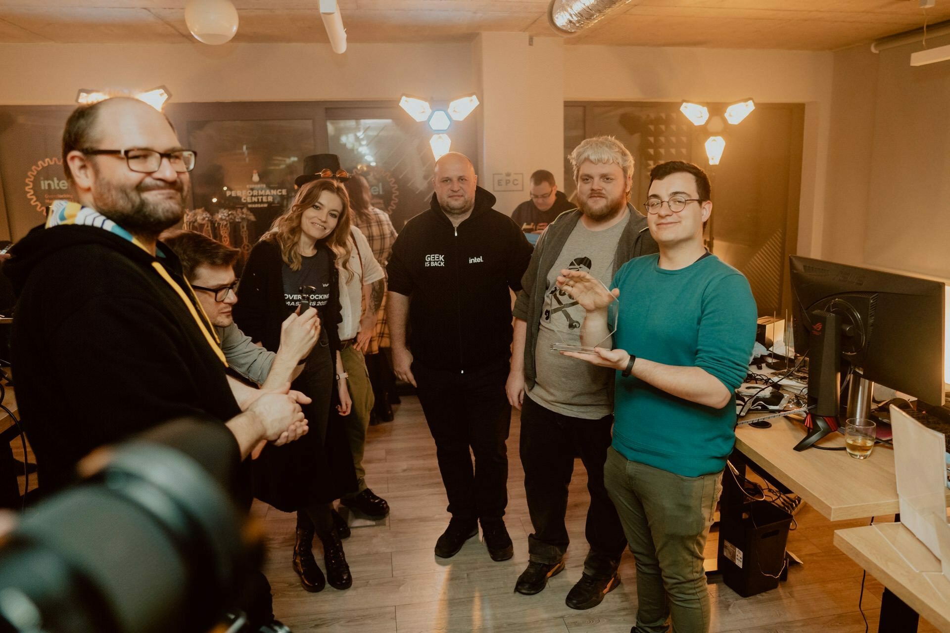 A group of people stand in an office space, smiling and posing for a photo. Some are holding objects in their hands. The room is illuminated by warm lighting fixtures, while in the background are desks, computers and various office supplies - perfect for an event photo shoot by event photographer Warsaw.  