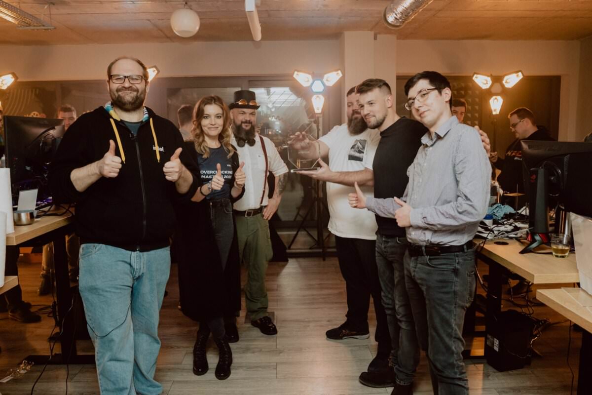 A group of six people stand in a room filled with computers and warm light, smiling and showing thumbs up. They seem relaxed and happy, with one person in the middle holding a small trophy. The room has a relaxed and friendly atmosphere, perfect for event photography.  