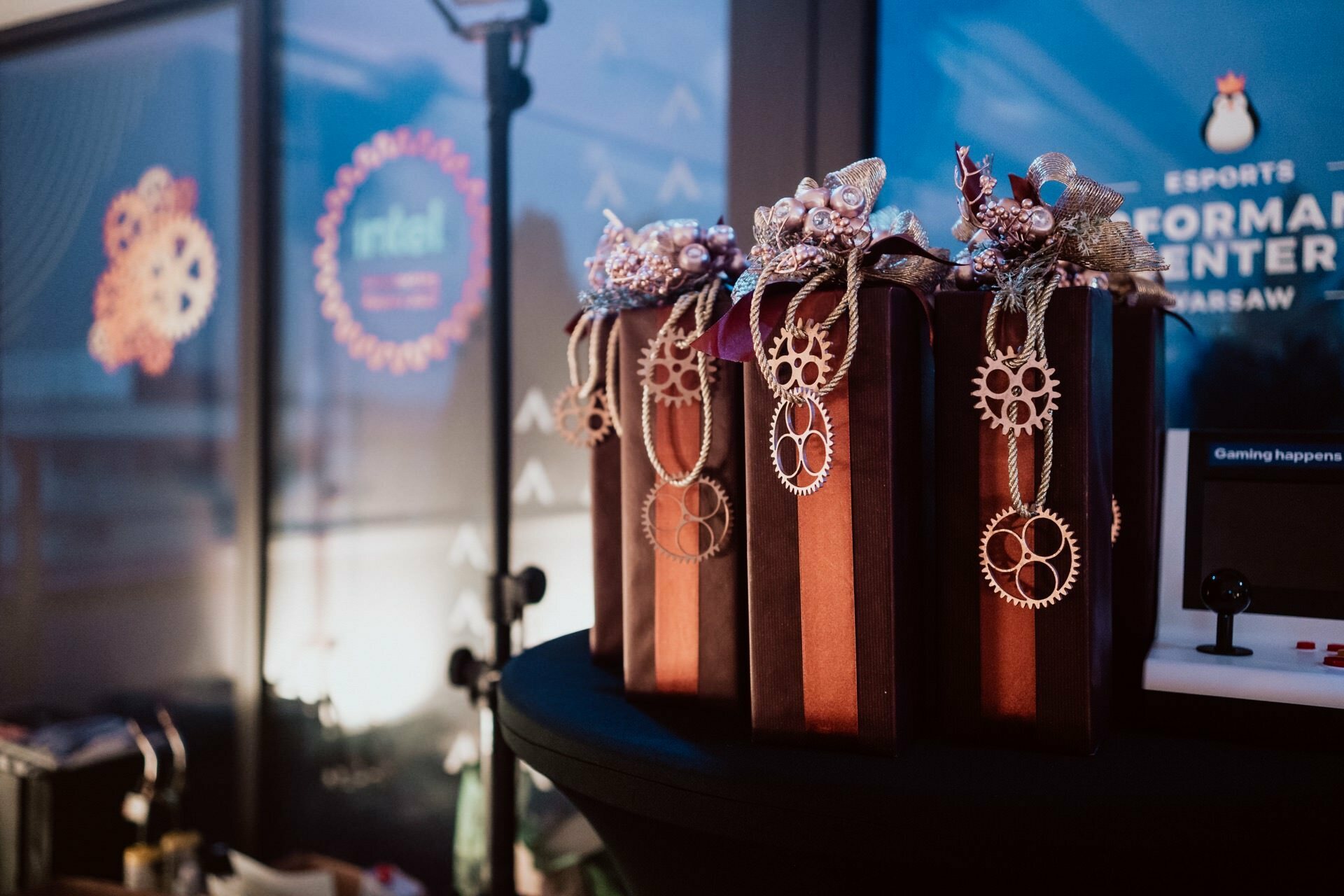 A display of prize boxes decorated with decorative pinions and ribbons, set on a table. In the background, a fuzzy banner with partially visible text and a soft focus on several light fixtures, suggesting a formal setting for the event - ideal material for an event photo essay. 