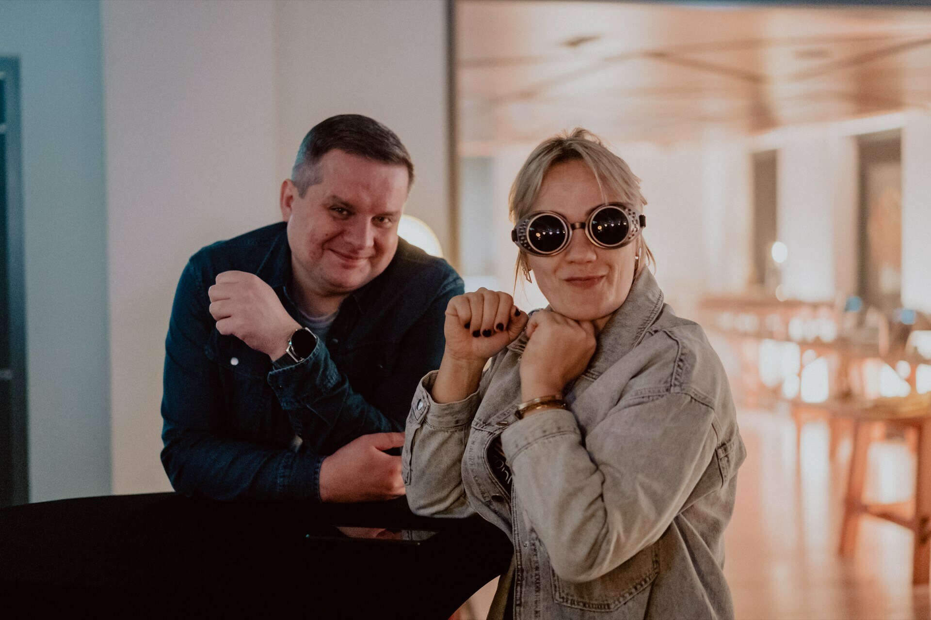 A man and a woman pose together in a casual setting in a room for an event photo. The man is wearing a denim shirt with a smartwatch on his wrist and rests his hand on a table. The woman is wearing a denim jacket and unique round sunglasses, adopting a playful pose with her fists raised.  