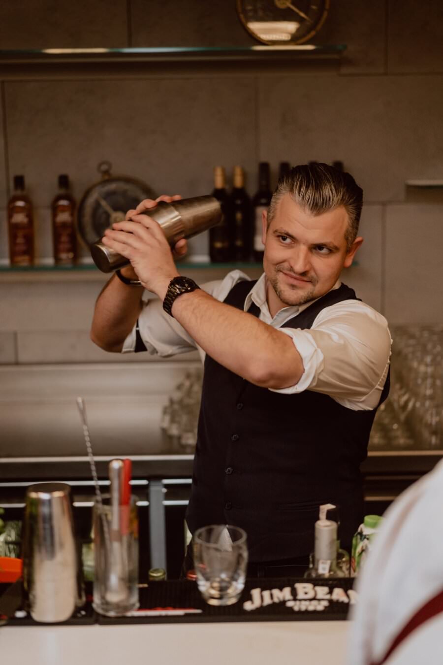 A bartender in a vest and white shirt shakes a shaker behind the bar counter. He is surrounded by liquor bottles and various bar tools, while glasses and a bar coaster labeled "Jim Beam" stand on the countertop. Captured by Marcin Krokowski, he smiles and focuses on making a drink during this event photo.  