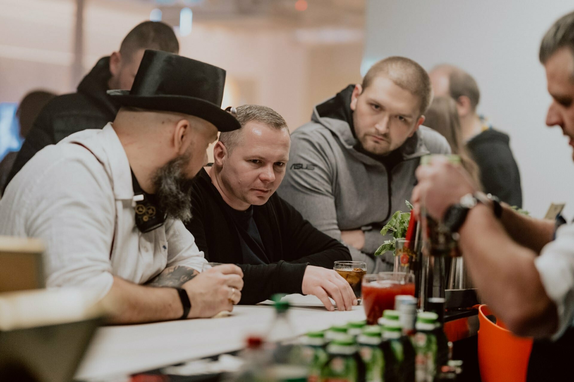 Three men sit at the bar while the bartender prepares drinks. One wears a cylinder and white shirt and chats with the others in casual attire. A variety of drinks and bar implements are visible on the counter, capturing a relaxed, sociable atmosphere, perfect for event photojournalism.  