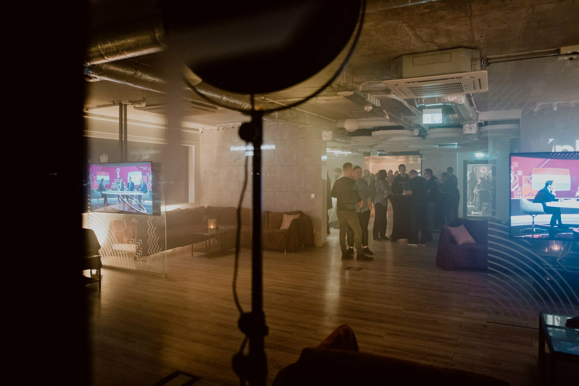 A dimly lit, modern indoor space with wooden floors and a cozy atmosphere. Groups of people mingle, some watching a screen displaying graphics and some people at an information desk. The scene, which is part of a photo essay of a Marcin Krokowski event, is viewed partly through a curtained, dark circular object.  