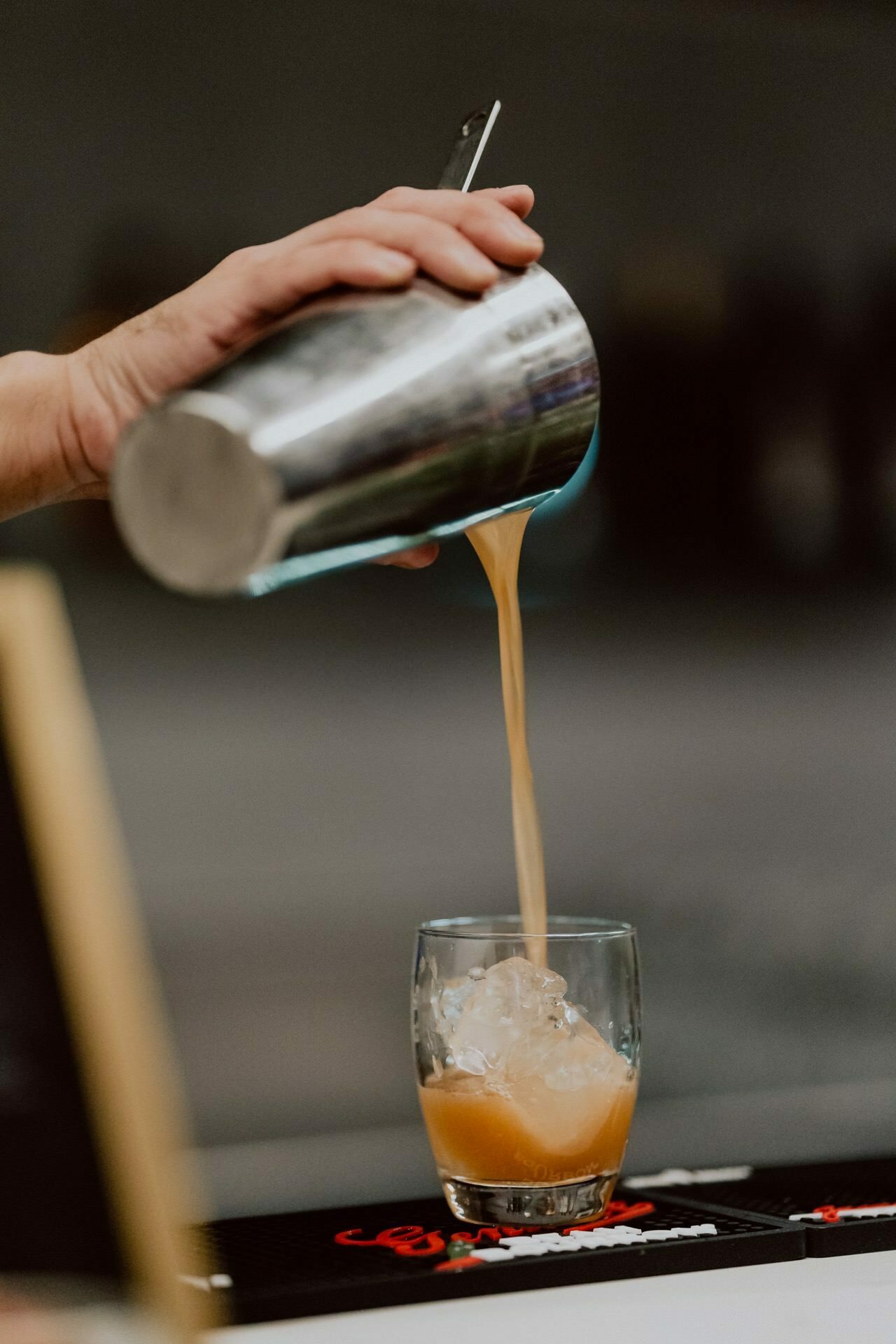 A person pours a mixed drink from a shaker into a glass filled with ice cubes standing on a bar top, showing the vibrant orange or reddish liquid of the fruity drink. The background is blurry, highlighting the pouring action, perfect for event photo coverage. 