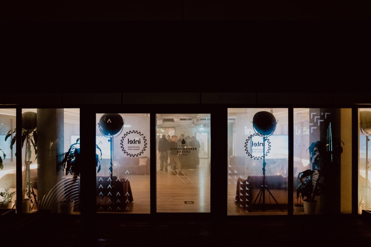 Night photo of a building with large glass doors and windows, lit from inside. The interior gives the impression of an office space where people can be seen. There are circular logos with intricate designs on the glass doors, captured as part of a photo essay of the event by Marcin Krokowski.  