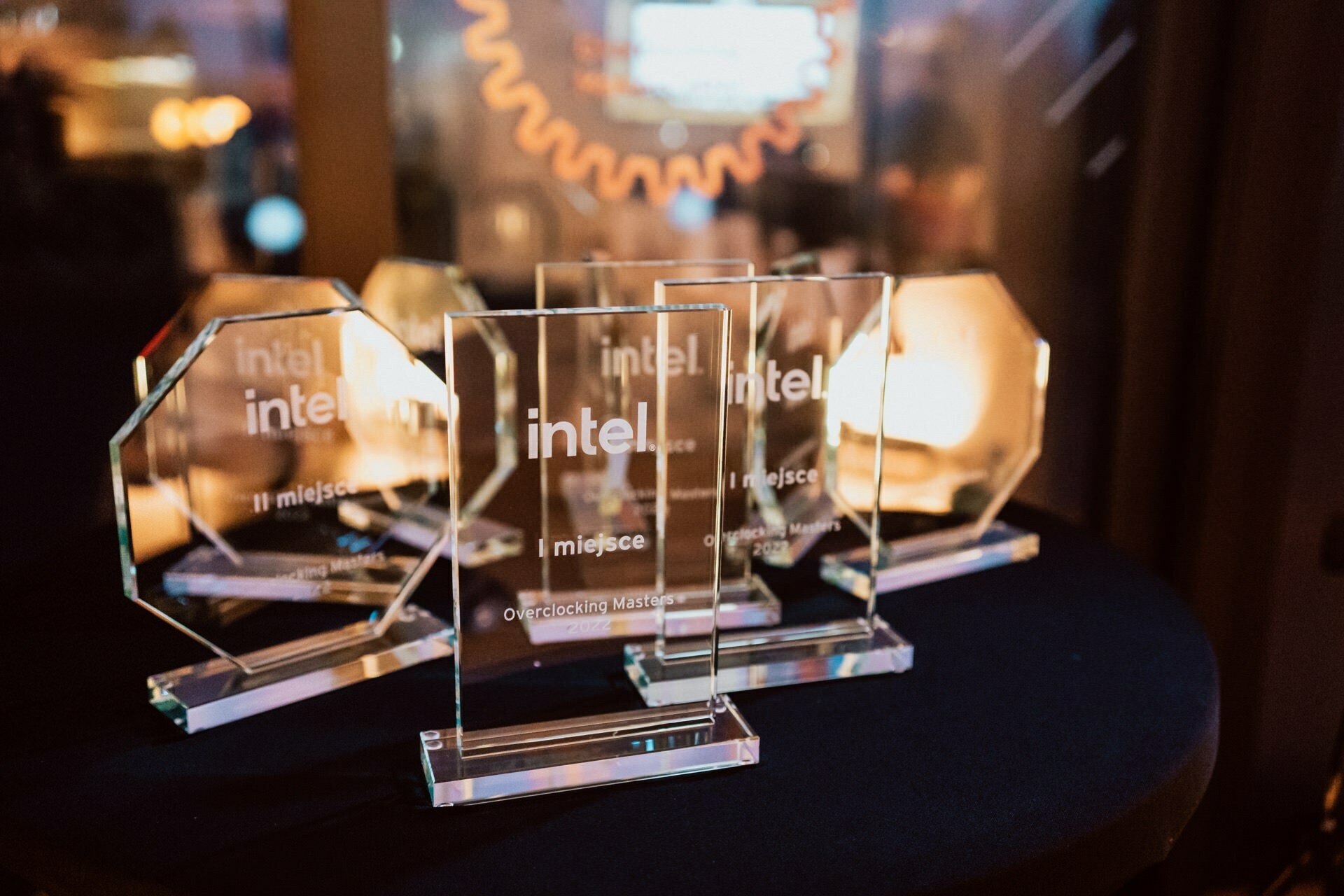 A group of acrylic awards with the Intel logo were displayed on a black table. On each award are inscriptions in Polish, with the words "1 place" (1st place) and "Champions of overclocking" in the center. The background is softly lit, creating a warm atmosphere, perfectly captured in this photo report of the event.  