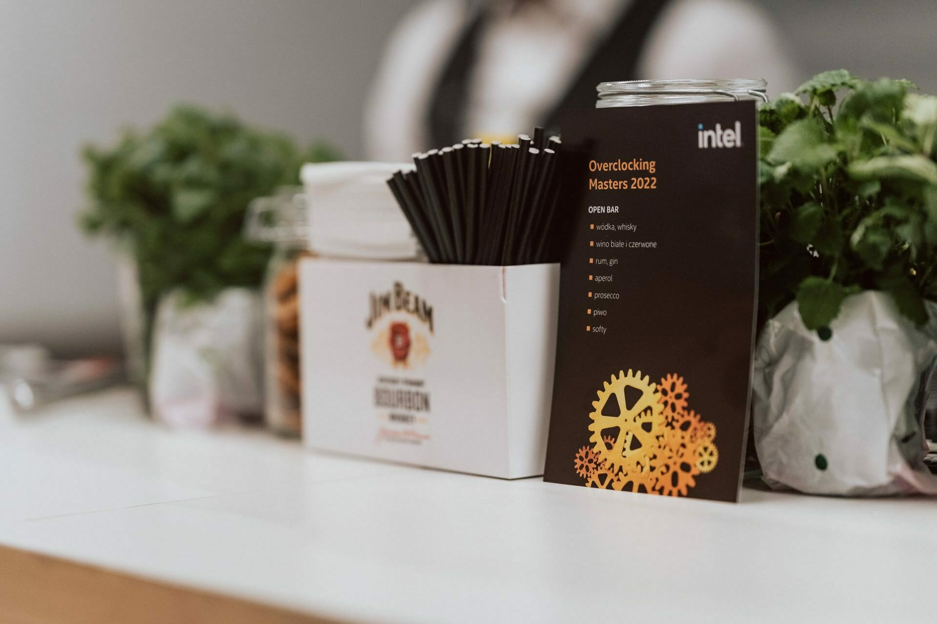 A counter with various items, including a box of whiskey, a black cup with straws and a black beverage menu titled "Overclocking Masters 2022" by Intel. The background is slightly blurred, capturing a person and green plants photographed by Marcin Krokowski. 