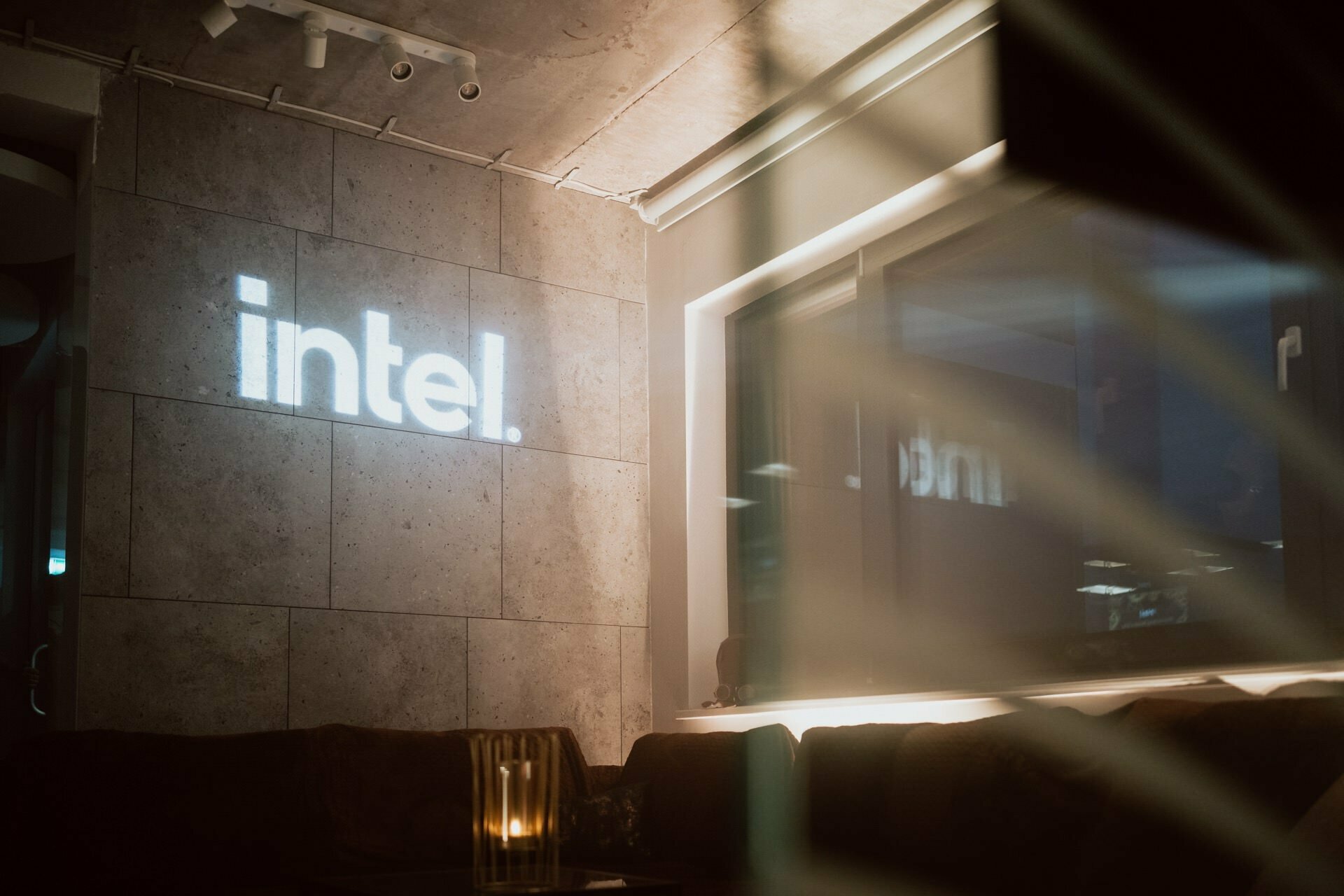 A dimly lit interior space with a concrete wall displaying an illuminated "Intel" logo. A candle on a table adds to the cozy atmosphere near the window reflecting the logo. The modern decor, highlighted in this event photo essay, includes a mix of soft and industrial elements.  