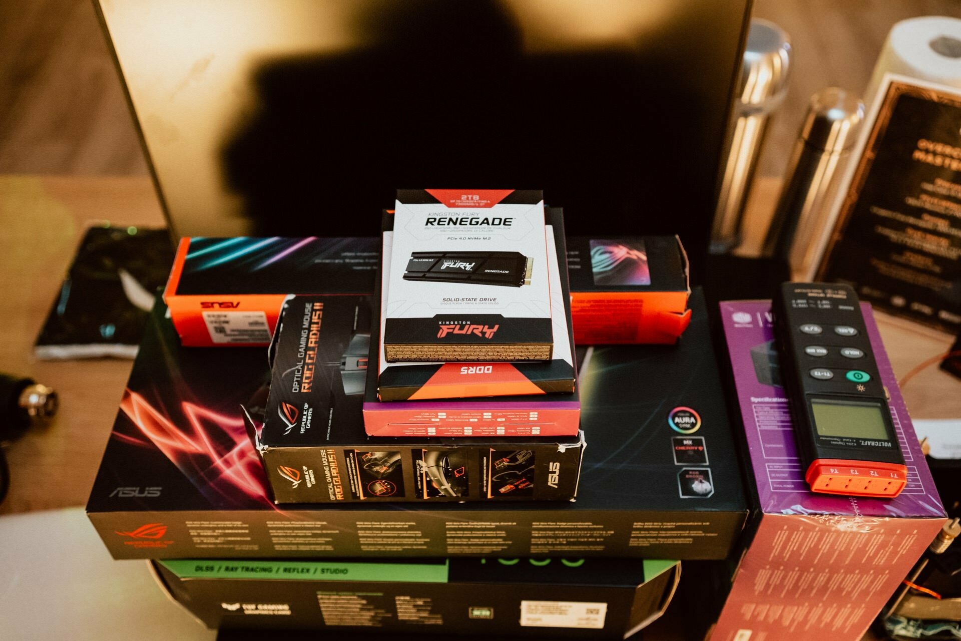 A stack of computer hardware components is stacked on the desk, including an SFX power supply, SSD, high-performance RAM and other peripherals from brands such as ASUS, Kingston Fury and ROG. A computer monitor stands in the background. The scene captures a moment worthy of Marcin Krokowski's event photography.  