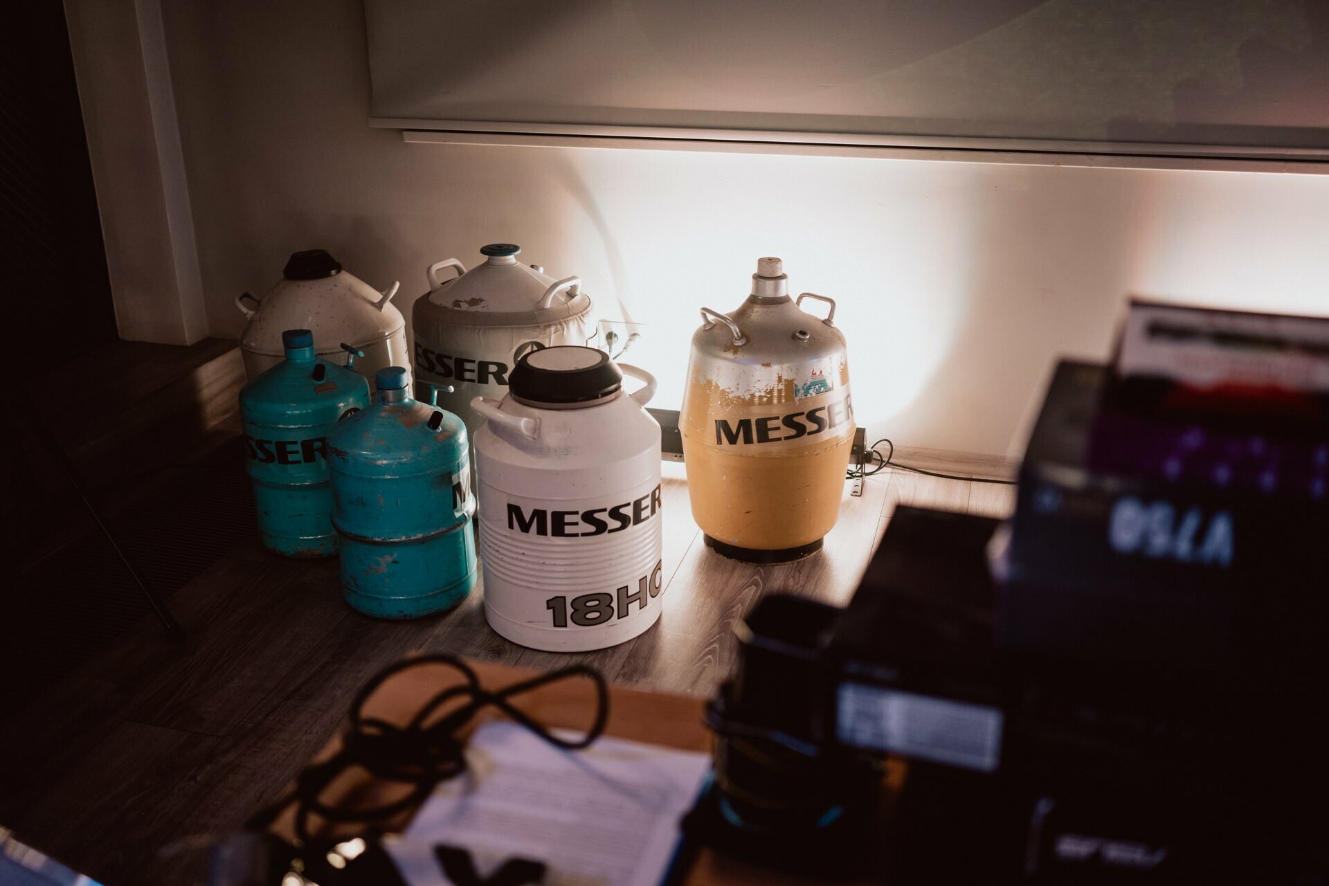 Several metal containers labeled "Messer" and "18H" of various sizes and colors were placed against a wall in a dimly lit room. Some are blue, while others are gray, white and yellow. In the foreground, various objects and cables can be seen on a table, capturing the essence of the event's photo coverage.  