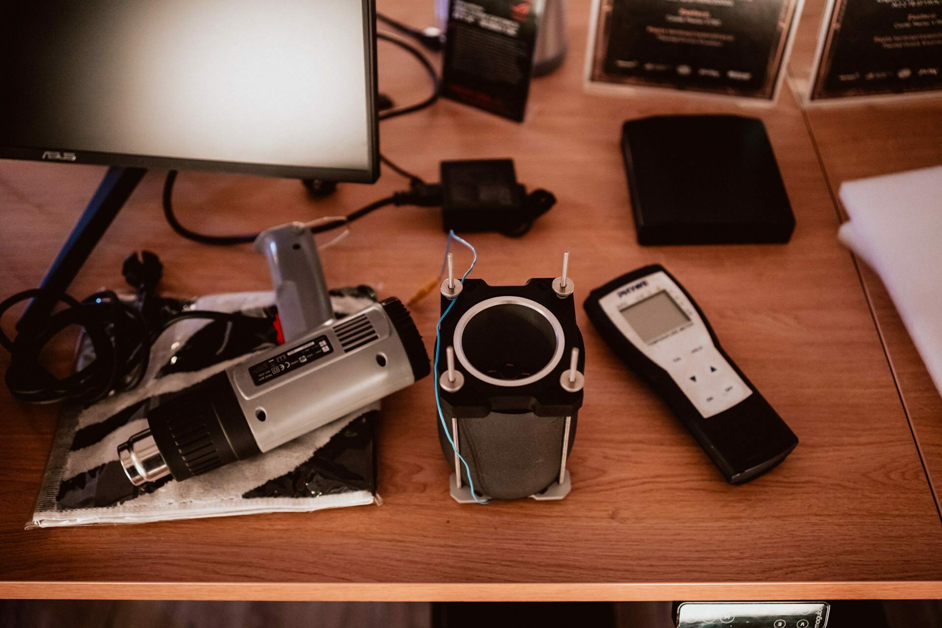 A wooden desk with a variety of electronic equipment, including a computer monitor, a tanner, a cylindrical mechanical device with wires, a handy electronic tester and an adapter. The items are neatly arranged on the desk, resembling preparations for photo-reporting events. 