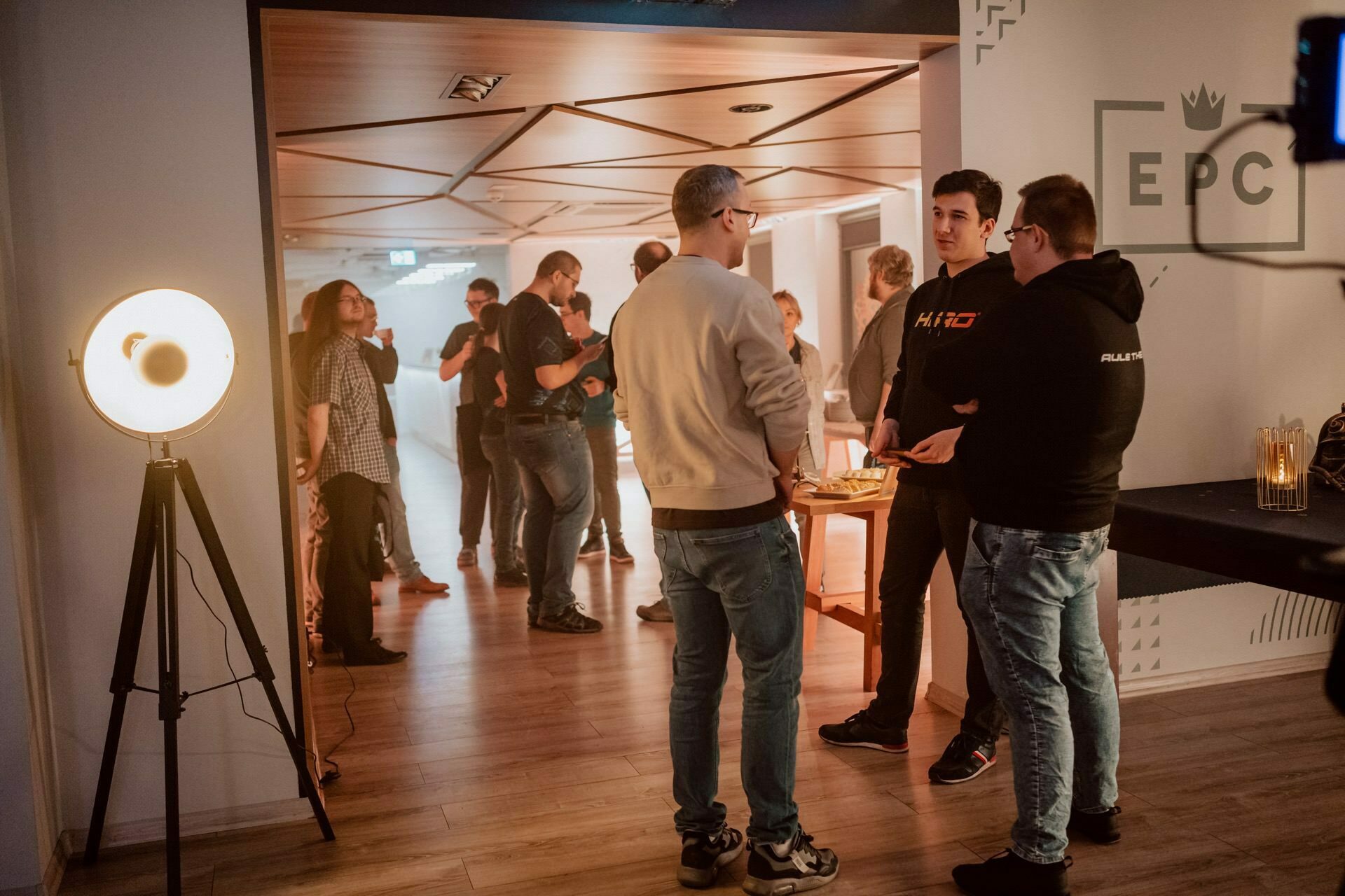 A group of people gather in a dimly lit room with a wooden floor and geometric ceiling. Three people are talking in the foreground, with other people mingling in the background. To the left is a tall floor lamp, which creates a warm atmosphere. On the right wall are the letters "EPC," capturing a striking event photography scene.   