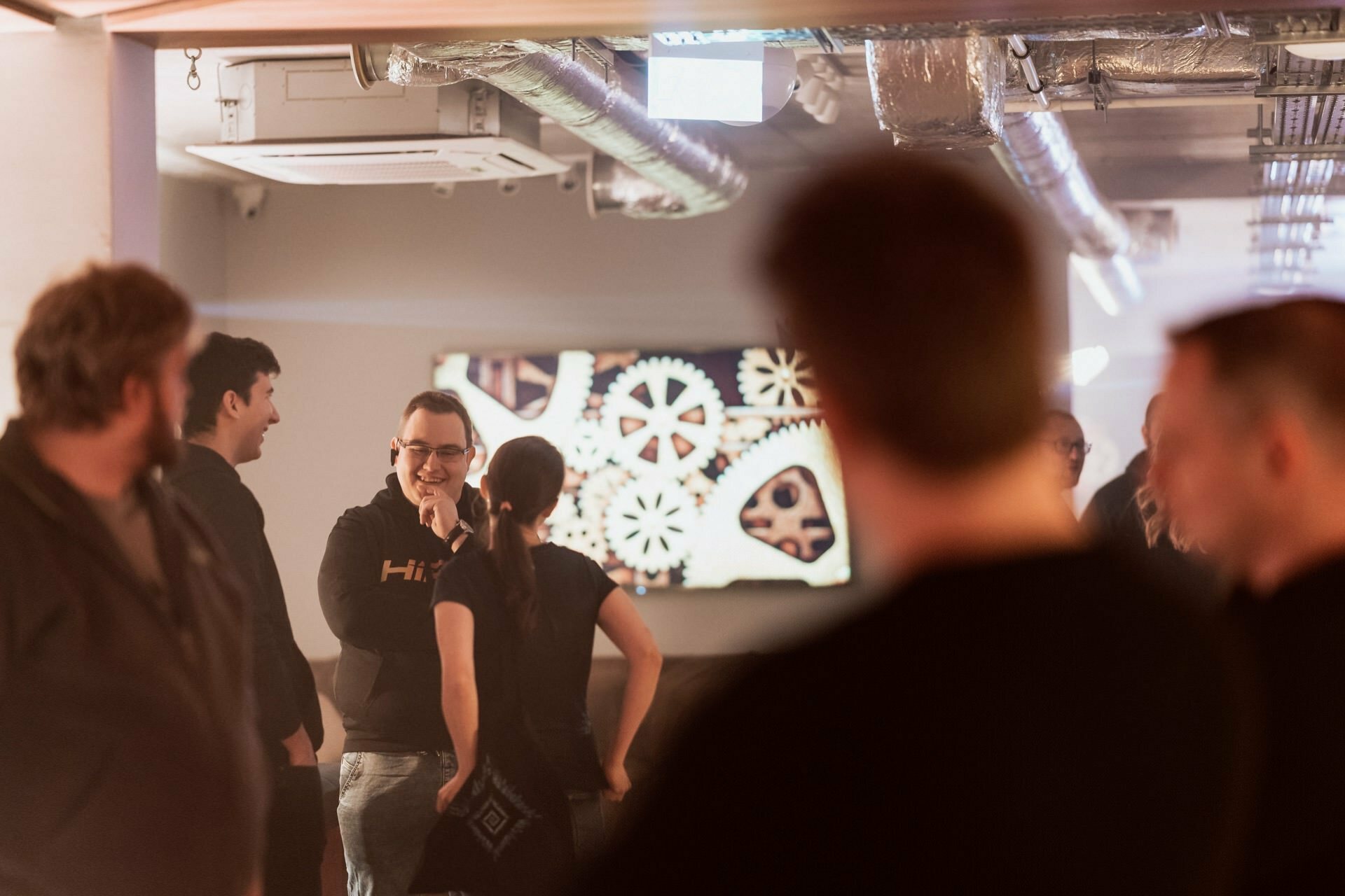 A group of people are in a room with industrial-style decor, equipped with a wall display showing interconnected gears. The relaxed atmosphere is beautifully captured in this event photography, showing two people smiling and talking to each other. 