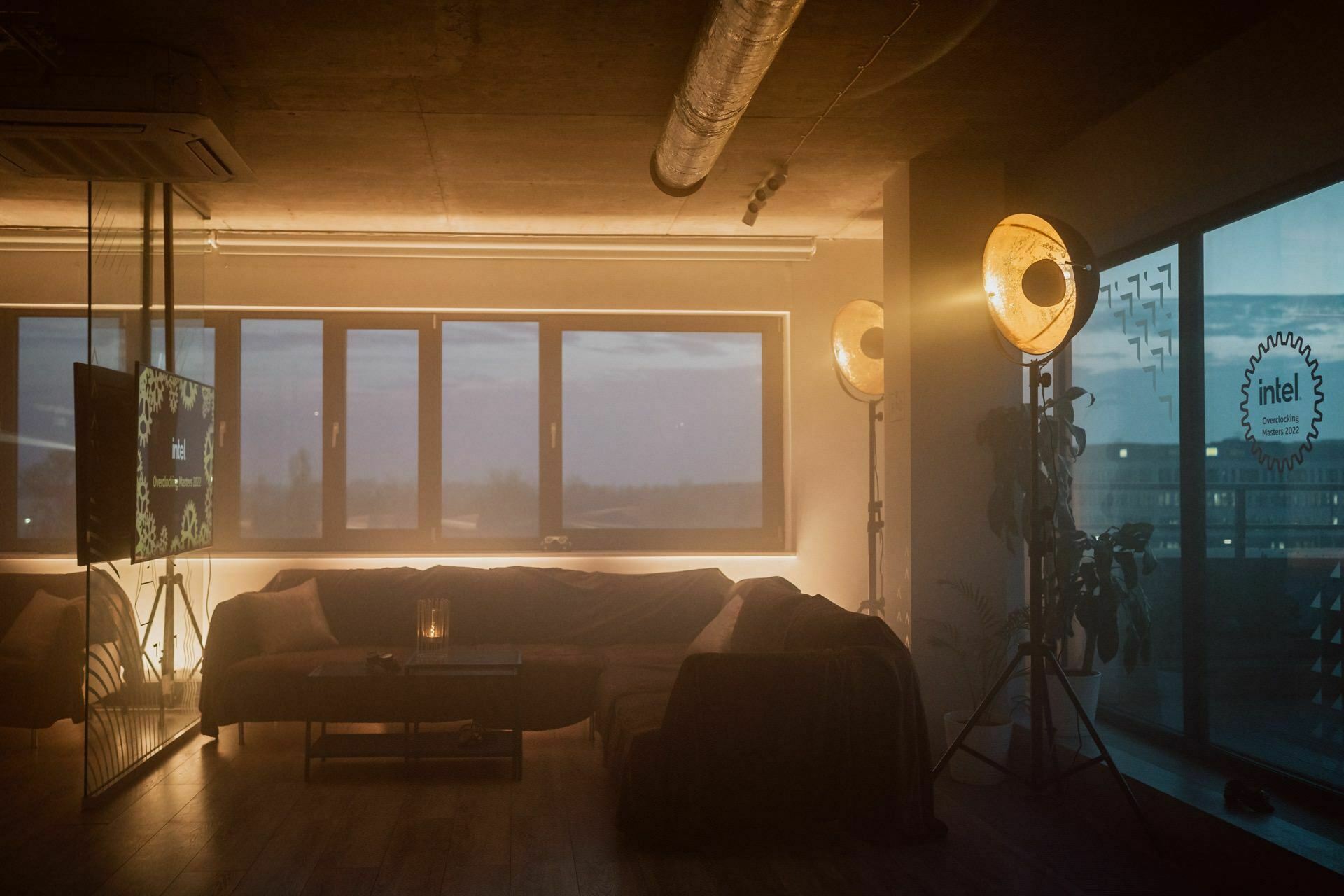 A cozy living room with a large L-shaped sofa, atmospheric lighting from standing lamps, a TV mounted on the wall and a coffee table. The room has large windows overlooking the city skyline at dusk. It's ideal for a party of friends or spending an evening with photo essays of events by Marcin Krokowski.  