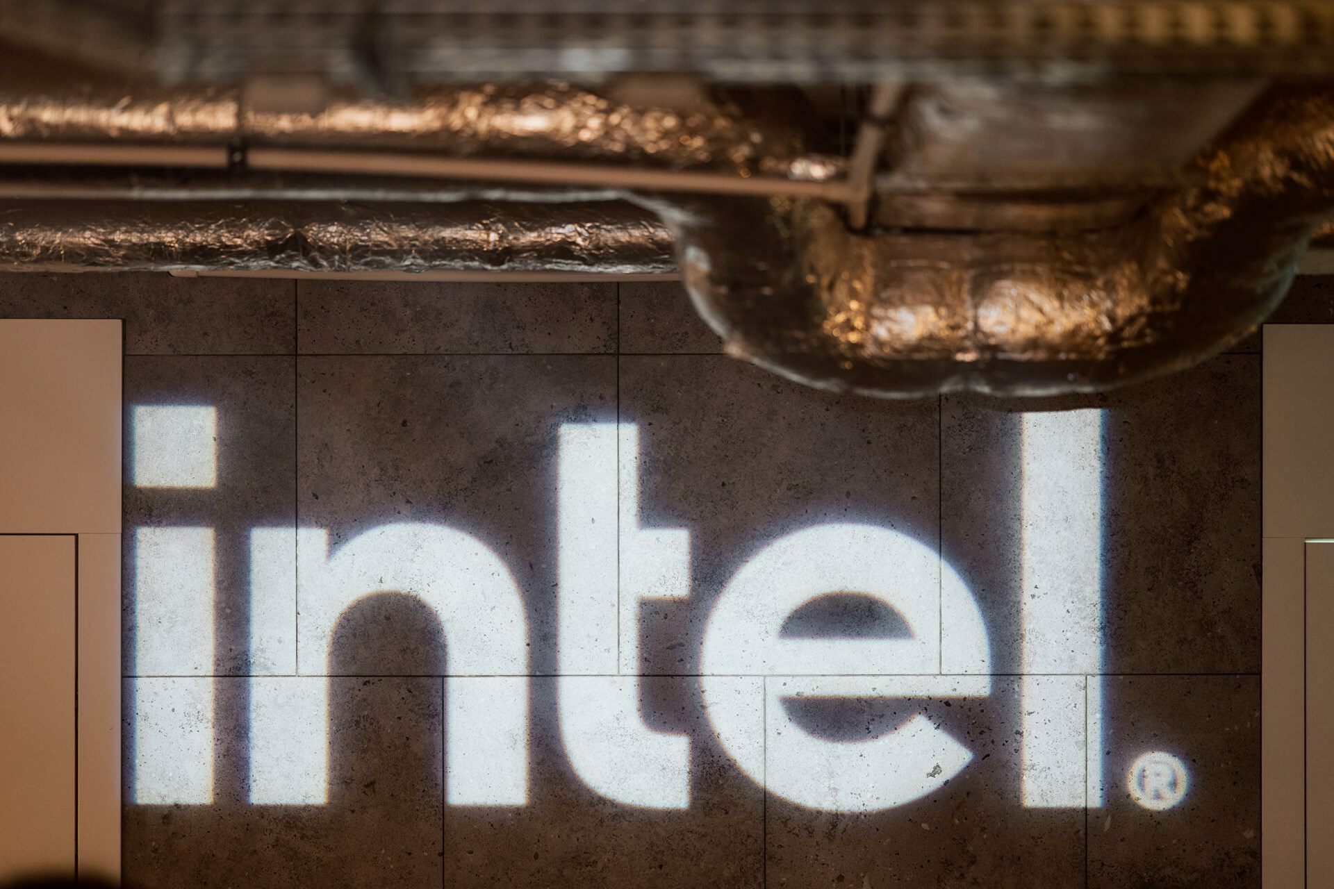 The photo shows the Intel logo displayed on a gray textured wall with exposed ducts running across the ceiling. The logo is clearly visible in white against a contrasting background, capturing a moment from a professional photoshoot of the event. 