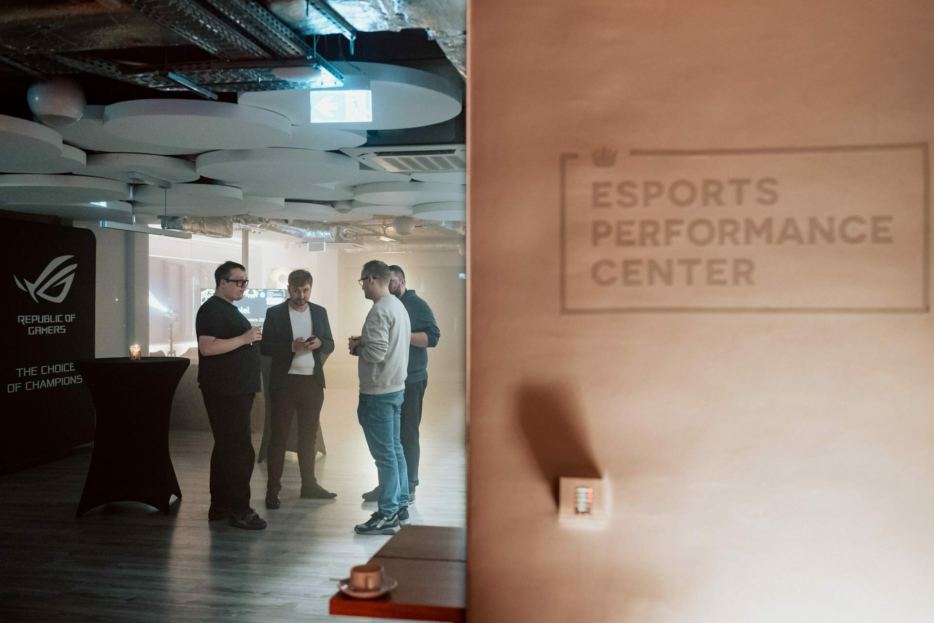 Four people stand and discuss in a modern room with overhead pipes and various lighting. A sign on the wall reads "Esports Performance Center," and a banner reading "Republic of Gamers" can be seen in the background, providing a backdrop for photo coverage of the events. 