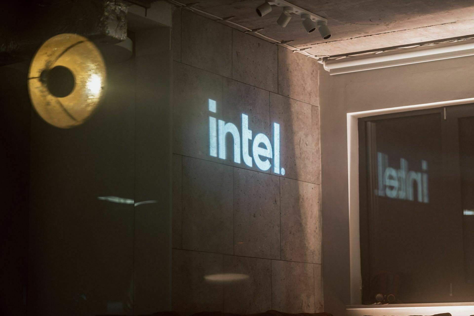 A dimly lit room with a concrete wall displaying the illuminated word "intel." The light projection is reflected in the window on the right. A circular lamp hangs near the ceiling, casting a yellowish glow on the wall, creating an atmosphere perfect for an event photographer warsaw session.  