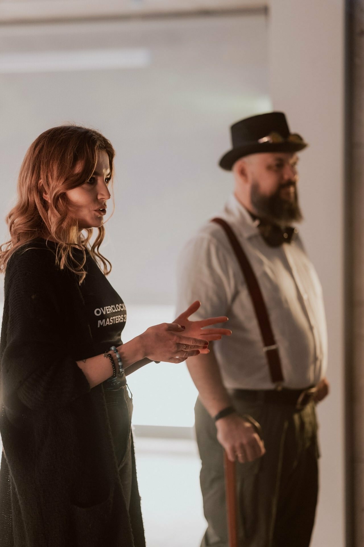 Says a woman with shoulder-length hair, gesturing with her hands. She is wearing a dark outfit. Next to her stands a bearded man wearing glasses, a shirt with suspenders and a hat. They appear to be indoors, probably giving a presentation or doing event photography.   