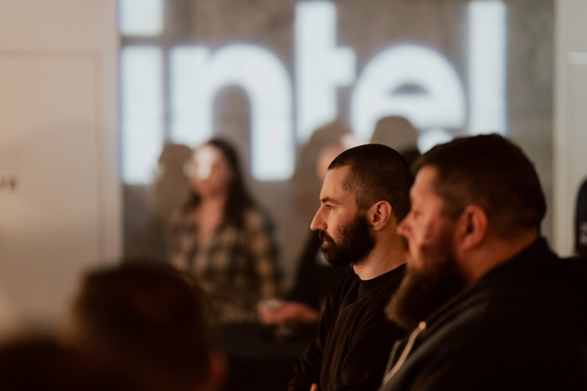 During the presentation, a group of people listen attentively. The word "Intel" is clearly highlighted in the background. Two bearded men stand in the foreground, focused on the speaker. In the blurry background you can see other participants in the event, captured in an immersive photo essay of the events by Marcin Krokowski.   