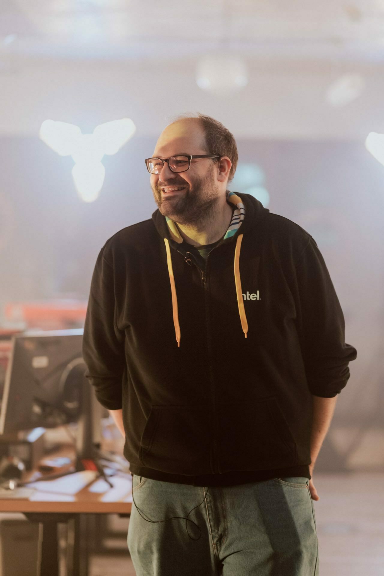 A bespectacled person wearing a black hoodie with "Intel" written on it smiles, standing in a room with his hands in his pockets. In the background there is soft lighting, a blurry computer monitor and furniture. This photo could successfully be included in Marcin Krokowski's event photo essay.  