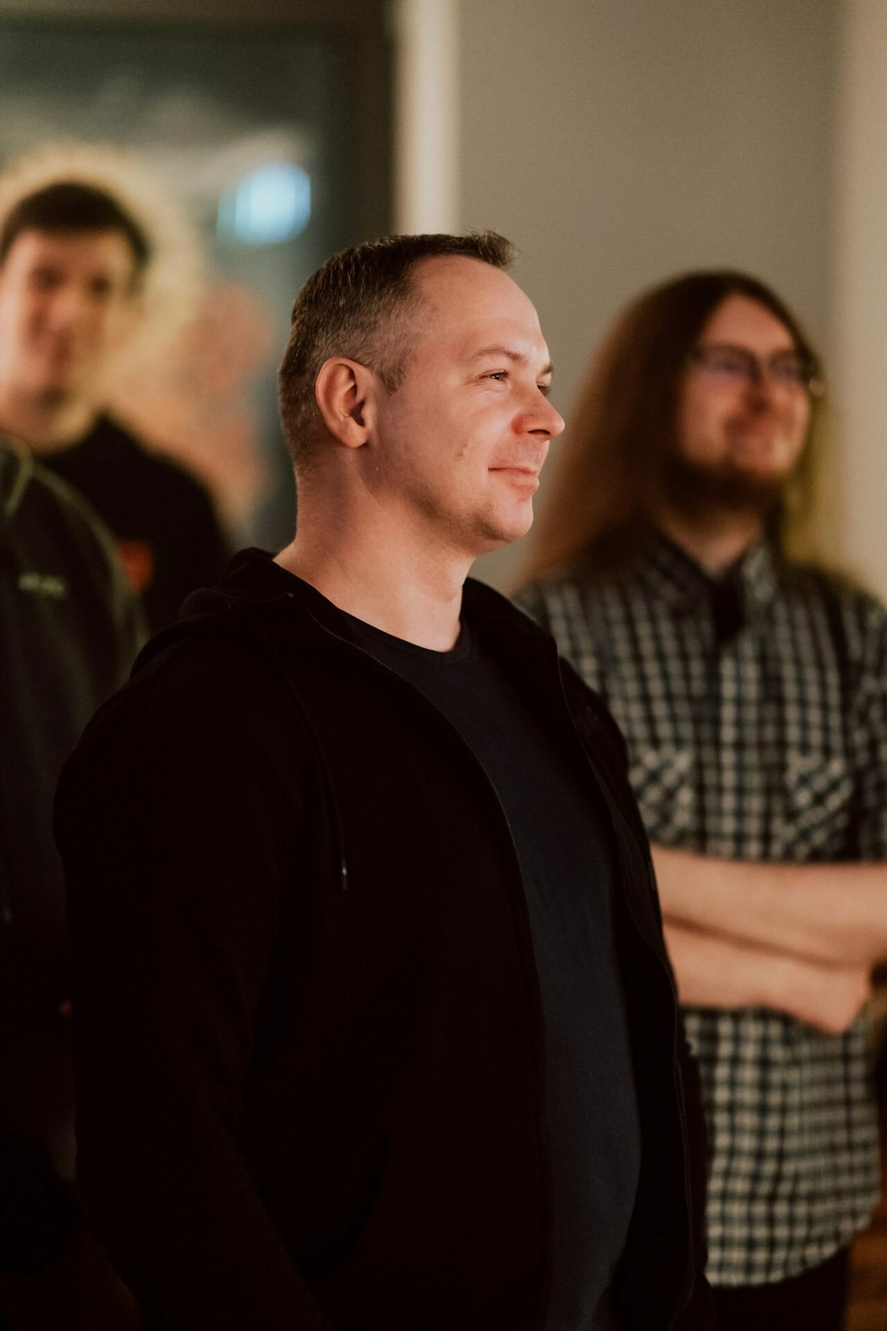 A man with short hair and a black hoodie smiles while standing with two other people, one with long hair and glasses, against a blurry background. The scenery appears to be in a room with soft lighting, which the event photographer captured perfectly. 