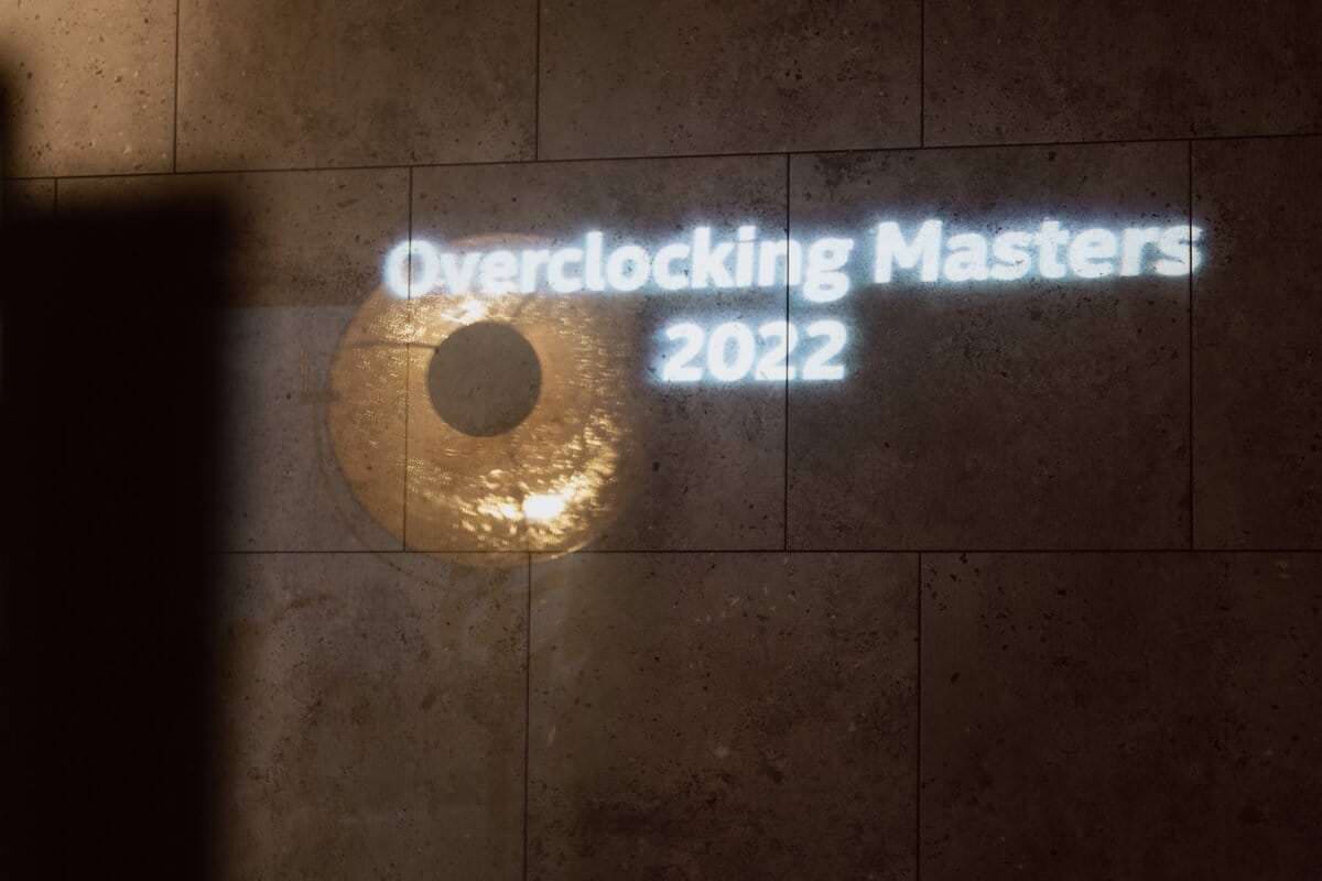  The displayed text reads "Overclocking Masters 2022" on a tiled wall, with a circular, illuminated object next to the text. The lighting creates a subtle shadow on the left side of the image, capturing the essence of event photography. 