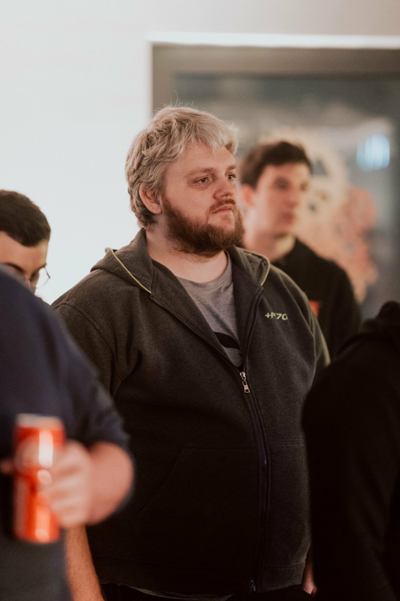 A person with light hair and beard stands attentively among a group of people. They are wearing a dark, zippered hooded sweatshirt, and the background is slightly blurred, suggesting a room setting. To the left of the image, another person is holding a can. This could be part of a photo essay of a Marcin Krokowski event.   