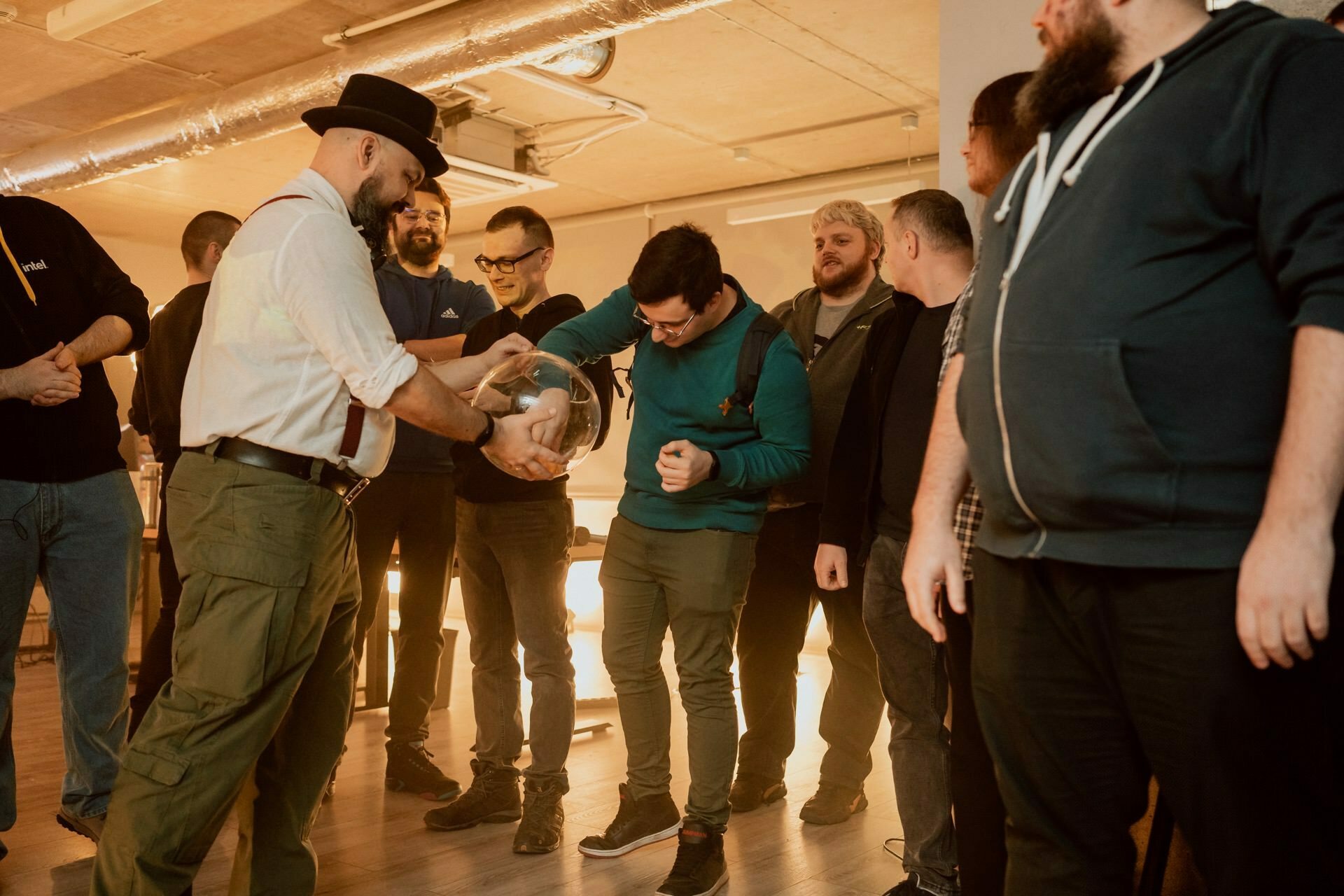 A group of men stand in a room, one of them is wearing a hat, white shirt and suspenders, holding a transparent bowl, and interacts with another man wearing glasses and a blue sweatshirt. The others gather around them and watch the interaction - a perfect moment for event photography. 