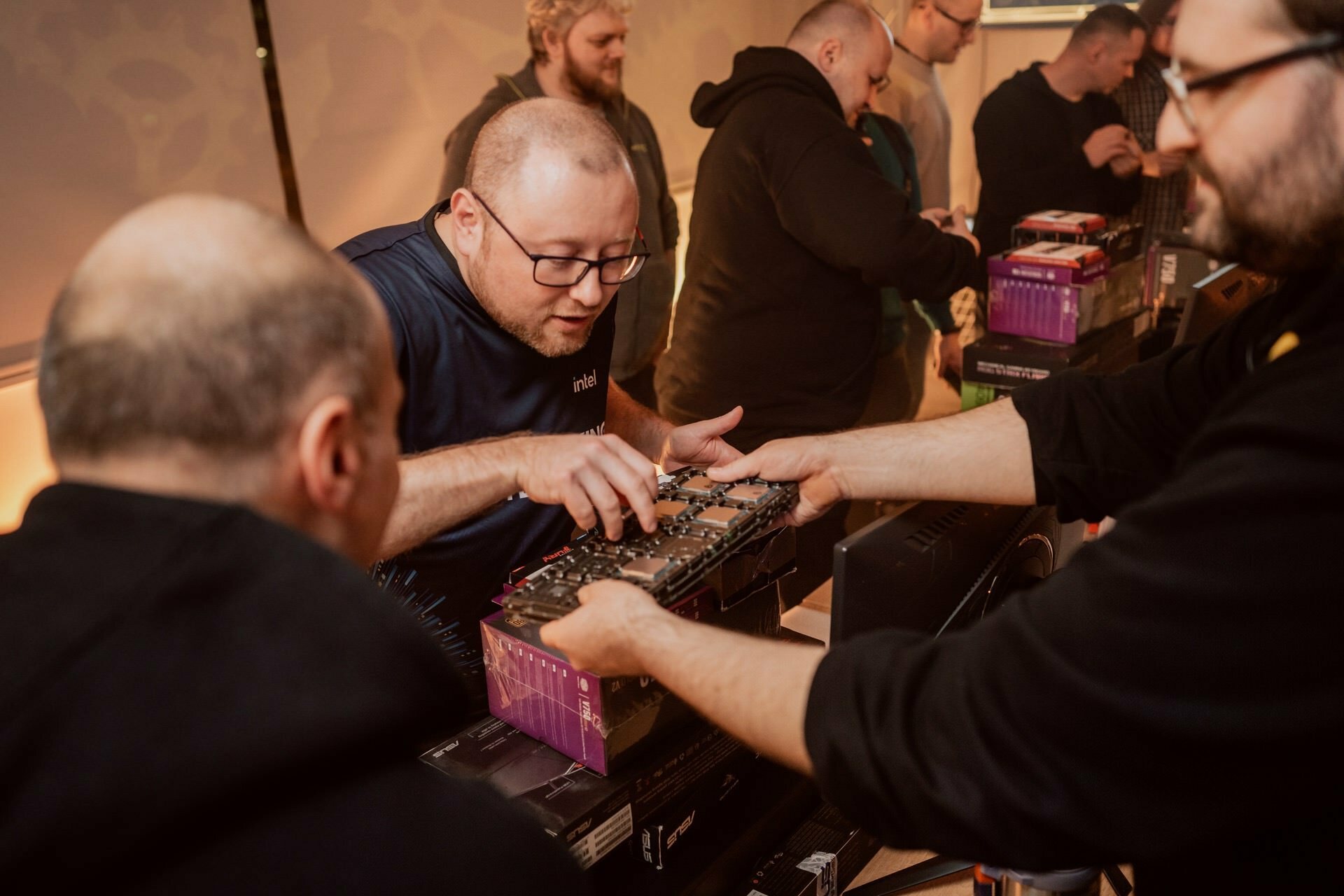 A group of people gather around a table and assemble computer components. One person in the middle carefully handles the motherboard, while the others help and observe. The room, captured in Marcin Krokowski's event photography, is warmly lit and the atmosphere is one of focus and cooperation.  