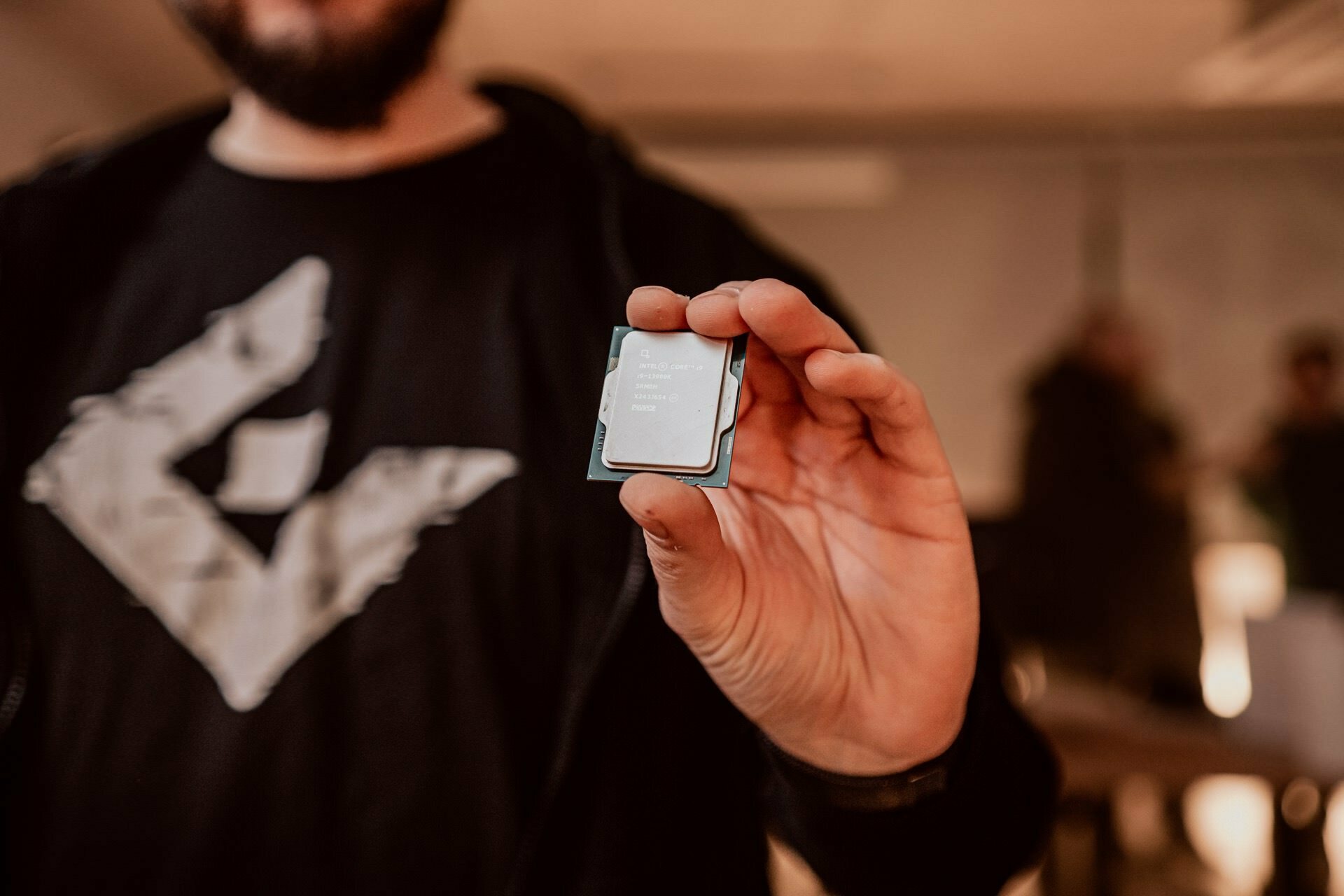 Marcin Krokowski, an event photographer, captures a person holding a small computer processor chip between his thumb and forefinger. The reflective surface of the chip shows text, and the person is wearing a black shirt and hoodie. The background is blurred with indistinct figures.  