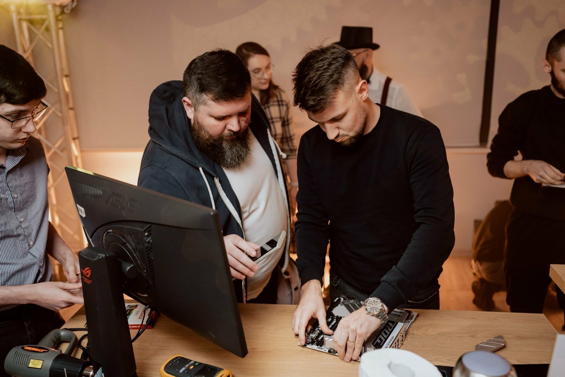 A group of five people have gathered around a wooden table, working on a disassembled computer. The man in the foreground meticulously handles the computer's components, while another man watches. A monitor and various tools can also be seen on the table, capturing the moment like a photo essay of the event.  