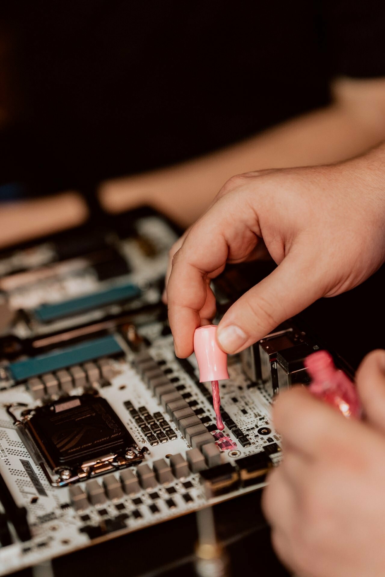 Close-up of a hand holding a brush with pink nail polish and applying it to a computer motherboard. The components and circuits of the motherboard are visible, and the other hand is partially visible in the background. This photo is part of Marcin Krokowski's unique photo essay of events.  