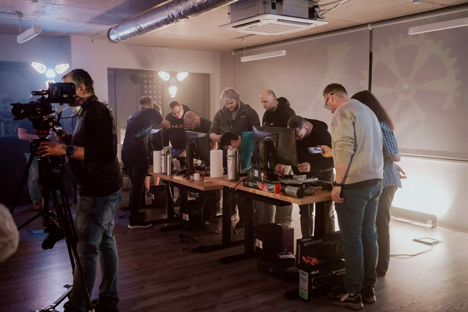 A group of people in a dimly lit room, some standing, some sitting, gather around tables with computer monitors. An event photographer from Warsaw captured the scene on the left, during which others are working on setting up computer equipment. 