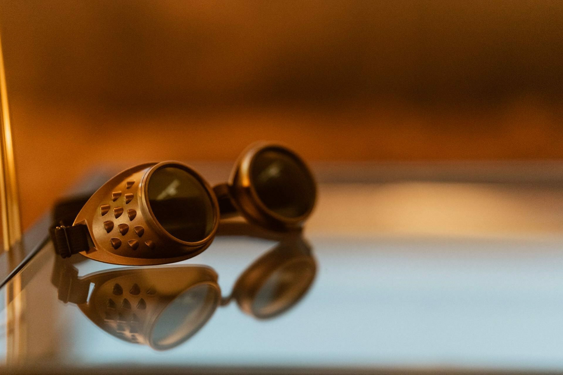 A pair of vintage-style aviator goggles with tinted lenses rest on a reflective surface, creating a clear reflection. The background is softly blurred with warm golden tones, giving the image a nostalgic feel, perfect for any event photography occasion. 