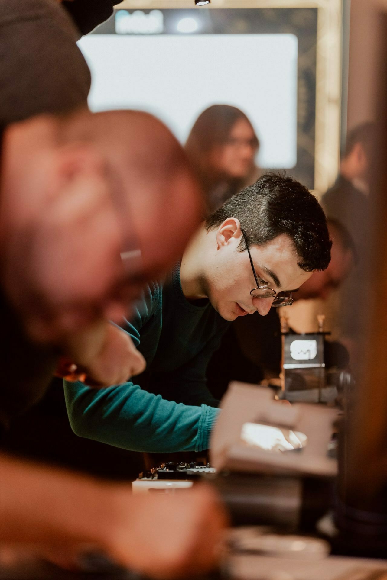 Marcin Krokowski, a man with glasses and short hair, is concentrating on an electronic device in a darkened room. Other people are blurred in the background, engaged in similar activities, creating a concentrated work environment. This scene captures the essence of a dedicated event photographer.  