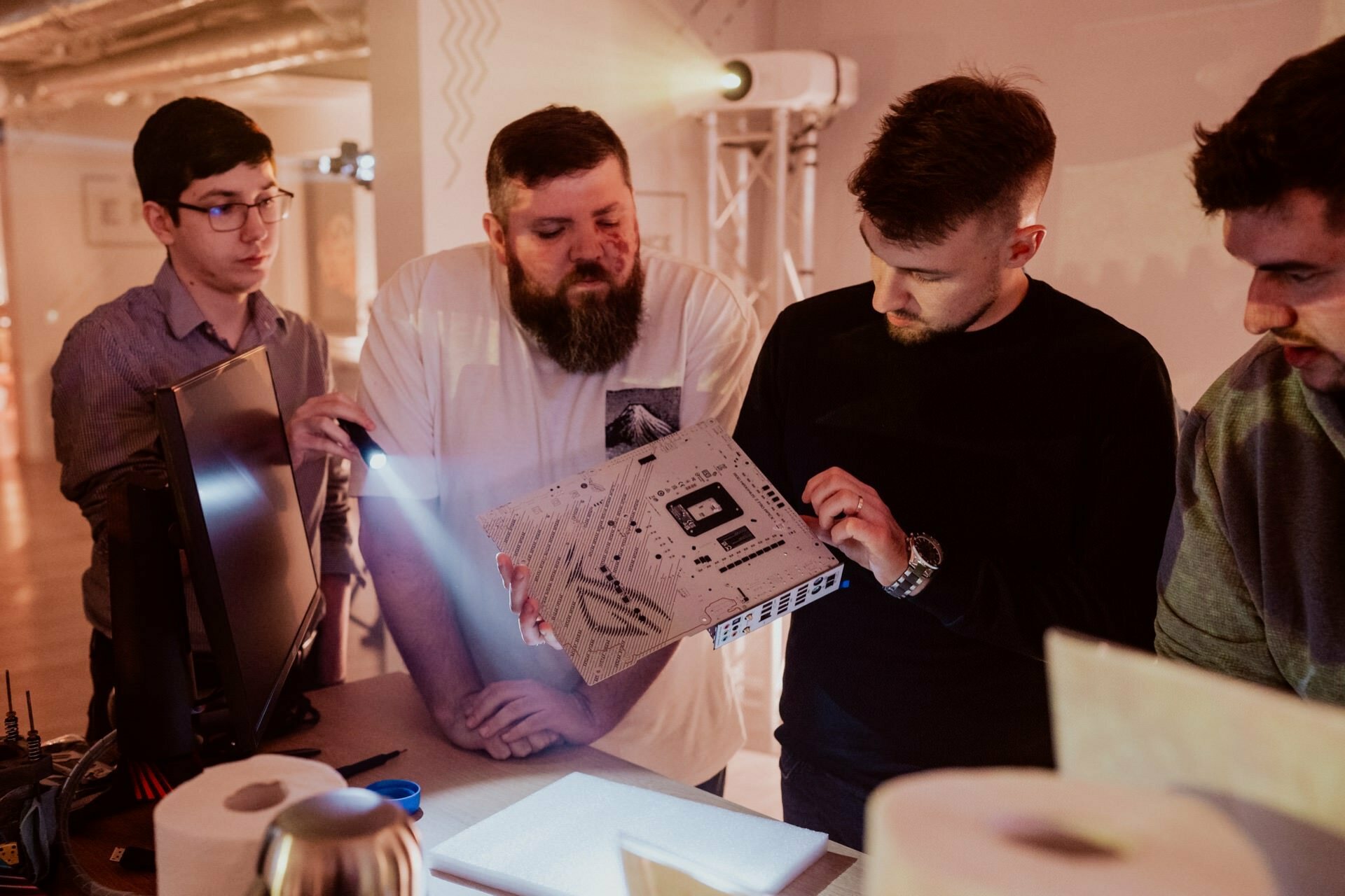 Four men gather around a table, examining some electronic equipment. One man is holding the device while another shines a light on it. They appear to be talking, probably troubleshooting or discussing its functions. A computer monitor is also visible in this event photo.   