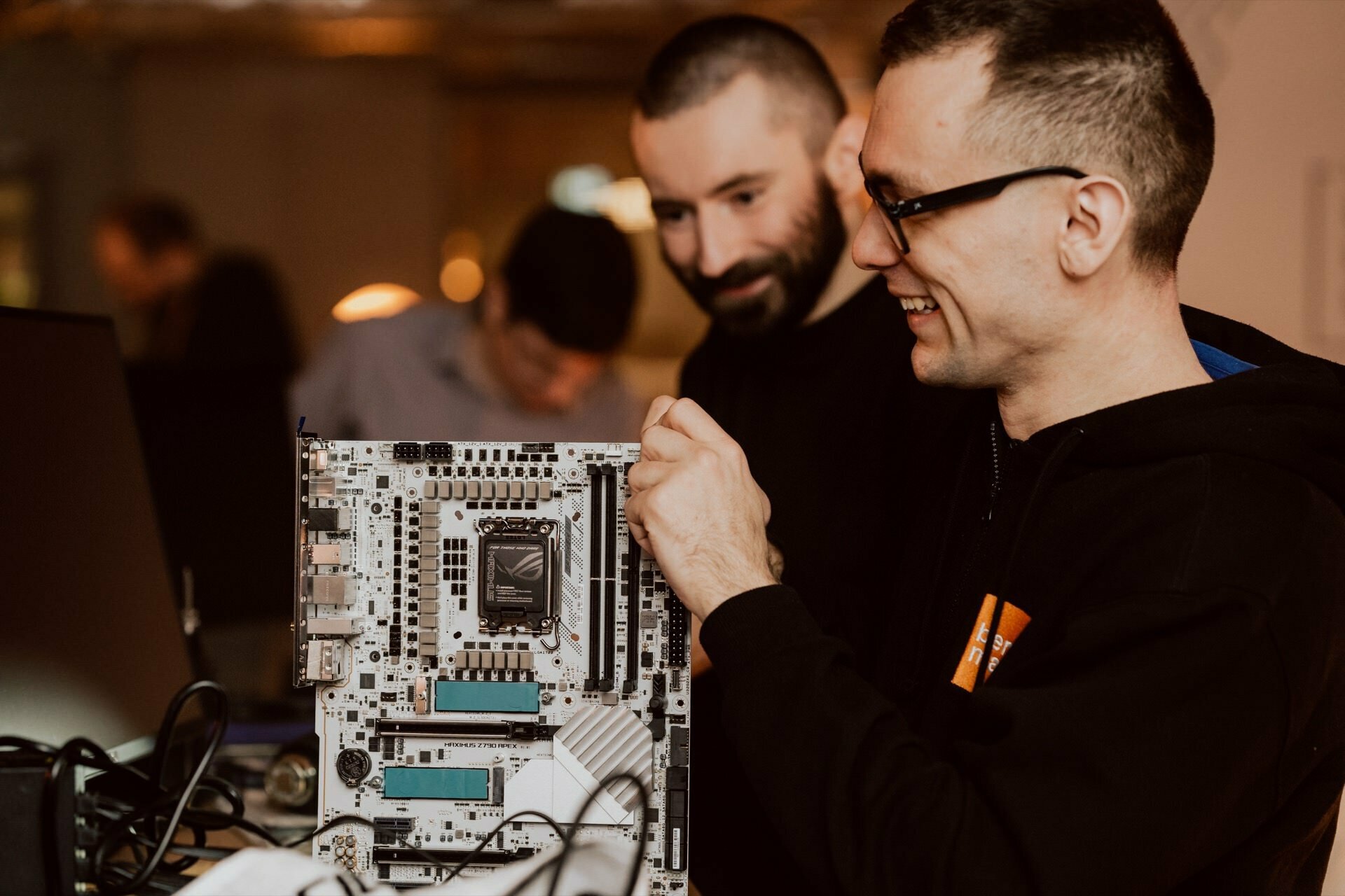 Two people are working together to assemble a computer. Both give the impression of being engaged and smiling. One person is holding and inspecting the motherboard, while the other is watching carefully. A blurry figure can be seen in the background capturing moments for a photo report of the event.   