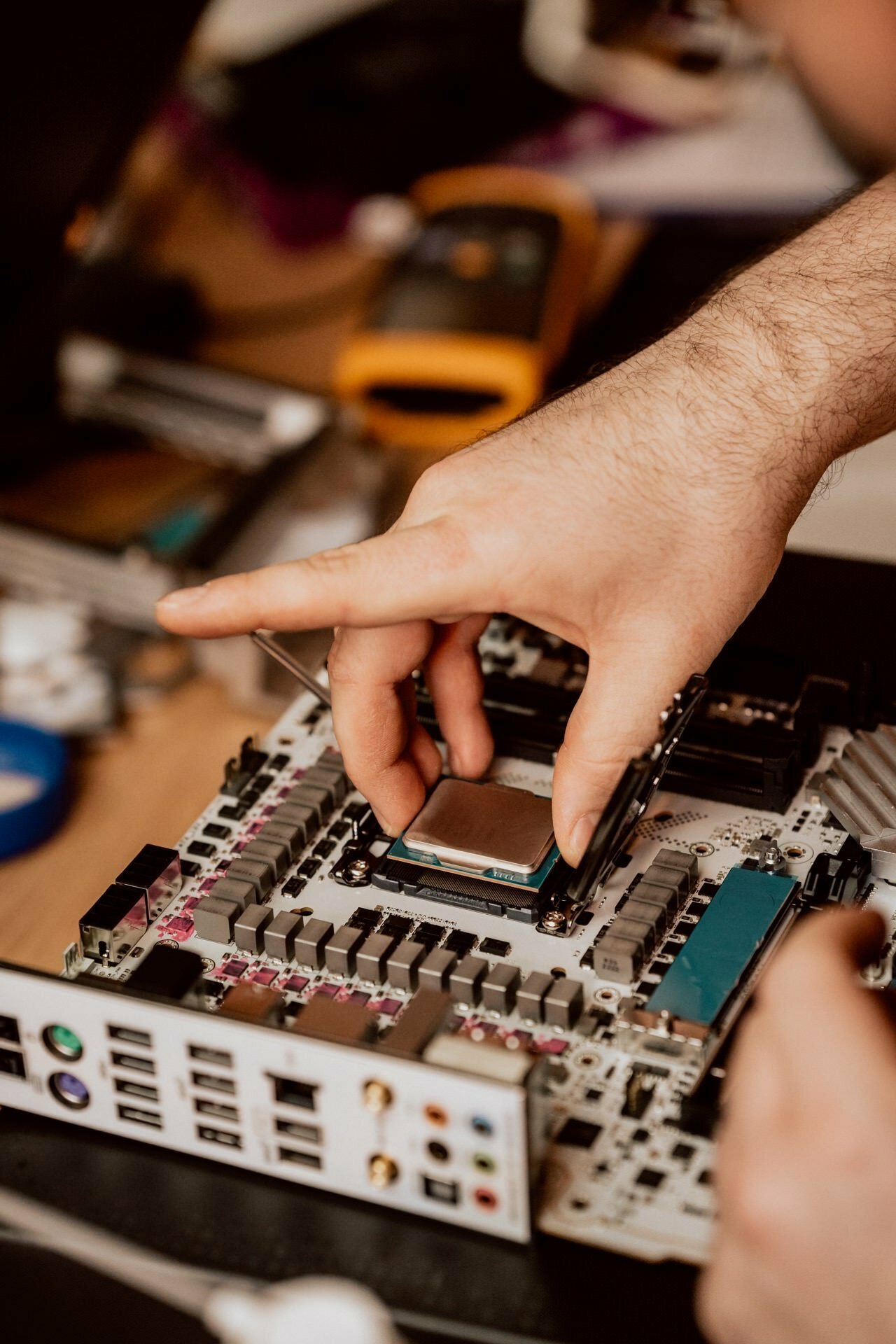 A man's hand installs a processor on a motherboard. The computer components are arranged on a table, and various ports and connections are visible on the motherboard. In the background are fuzzy electronic tools and devices, showing careful work along the lines of a photo-report of events.  