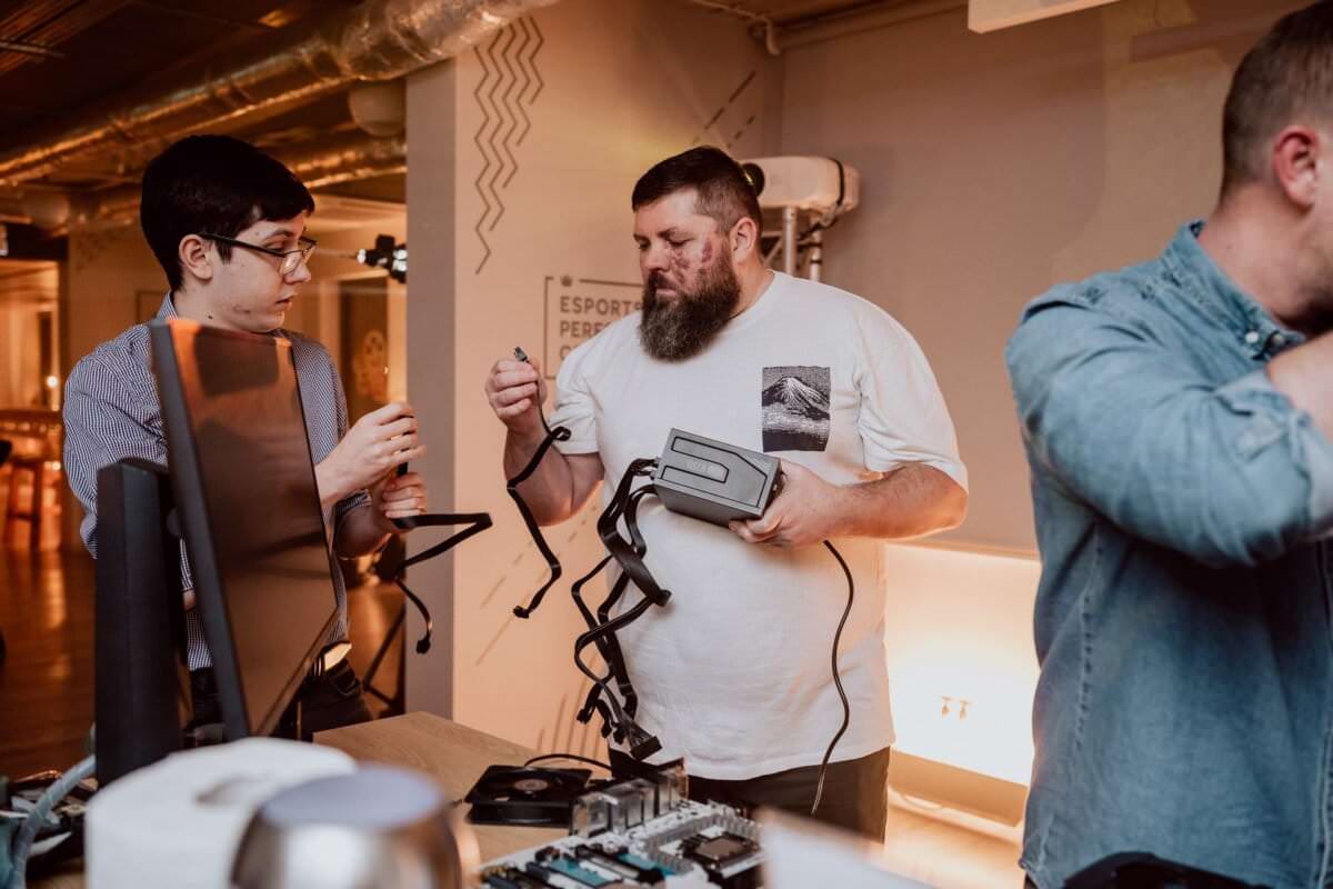 Two men sit at a table and configure or repair electronic equipment. One man in a white t-shirt is holding a power supply with cables, the other in a striped shirt is helping him. This scene is part of a photo essay of the event captured by an event photographer warsaw. The third person is partially visible on the right.   