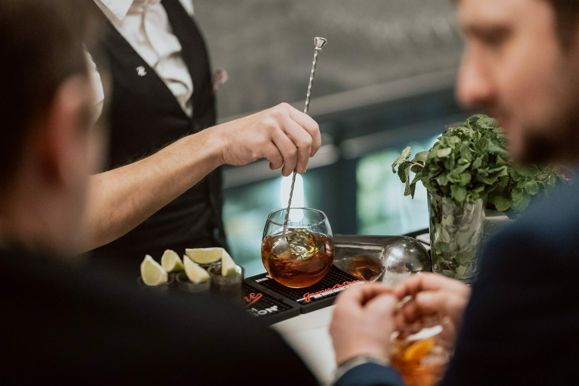 The bartender mixes a cocktail with a metal stirrer at the bar while two customers, seen from the shoulders up, carry on a conversation. On the countertop stand pieces of lime and a jar of fresh mint. This candid moment captures the essence of Marcin Krokowski's photojournalism.  