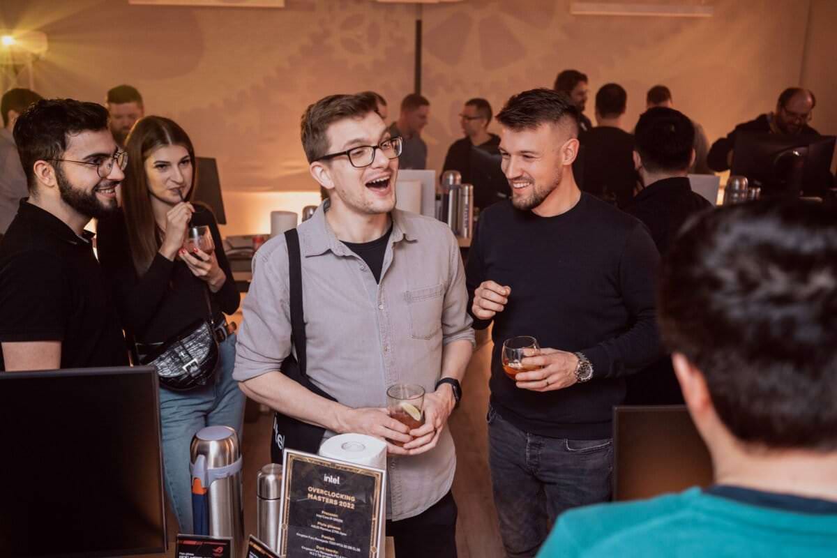 A group of people meet socially at a party held in a closed room. Two men in the middle are smiling and holding drinks, while others carry on a conversation or sip drinks. The atmosphere seems casual and friendly, with computer monitors and event signage visible in the background. This photo report of the events captures the lively mood of the meeting.   