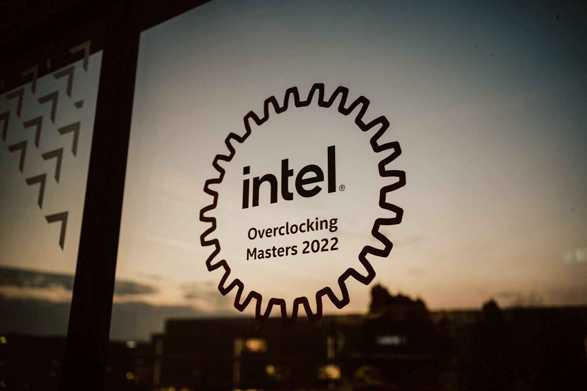 A large pinwheel-shaped sticker on the glass window shows the Intel logo with the text "Overclocking Masters 2022." In the background outside the window you can see the twilight sky and the silhouettes of buildings and trees, beautifully captured by Marcin Krokowski as part of his photo report of the event. 