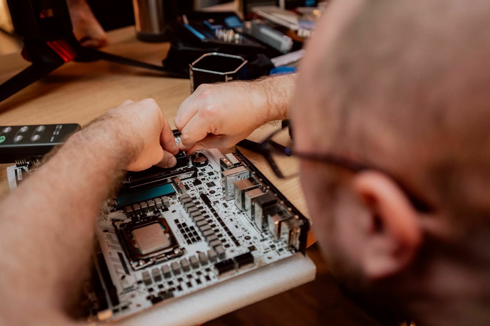 A person is assembling or repairing a computer motherboard, and his hands are diligently working on circuits. Various computer components and tools are visible on a wooden table. A person wearing glasses and looking focused can take a photo essay of events demonstrating technical skills.  